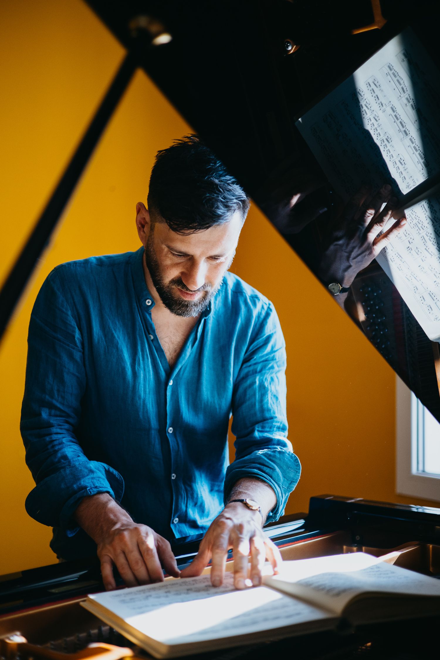 Bearded man in blue linen shirt reading sheet music at a grand piano against yellow wall.