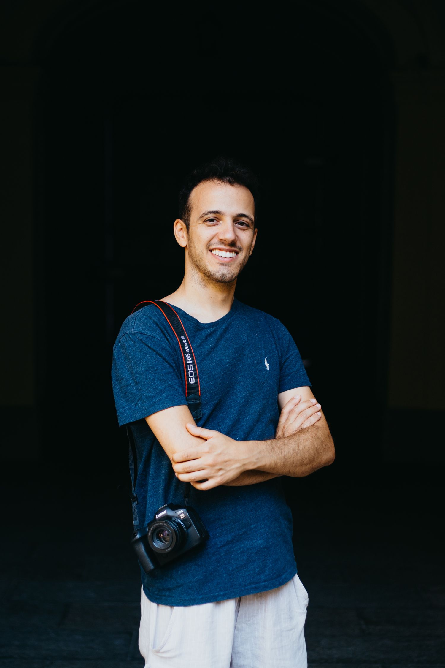 Smiling young male photographer with Canon camera poses with arms crossed against dark background.
