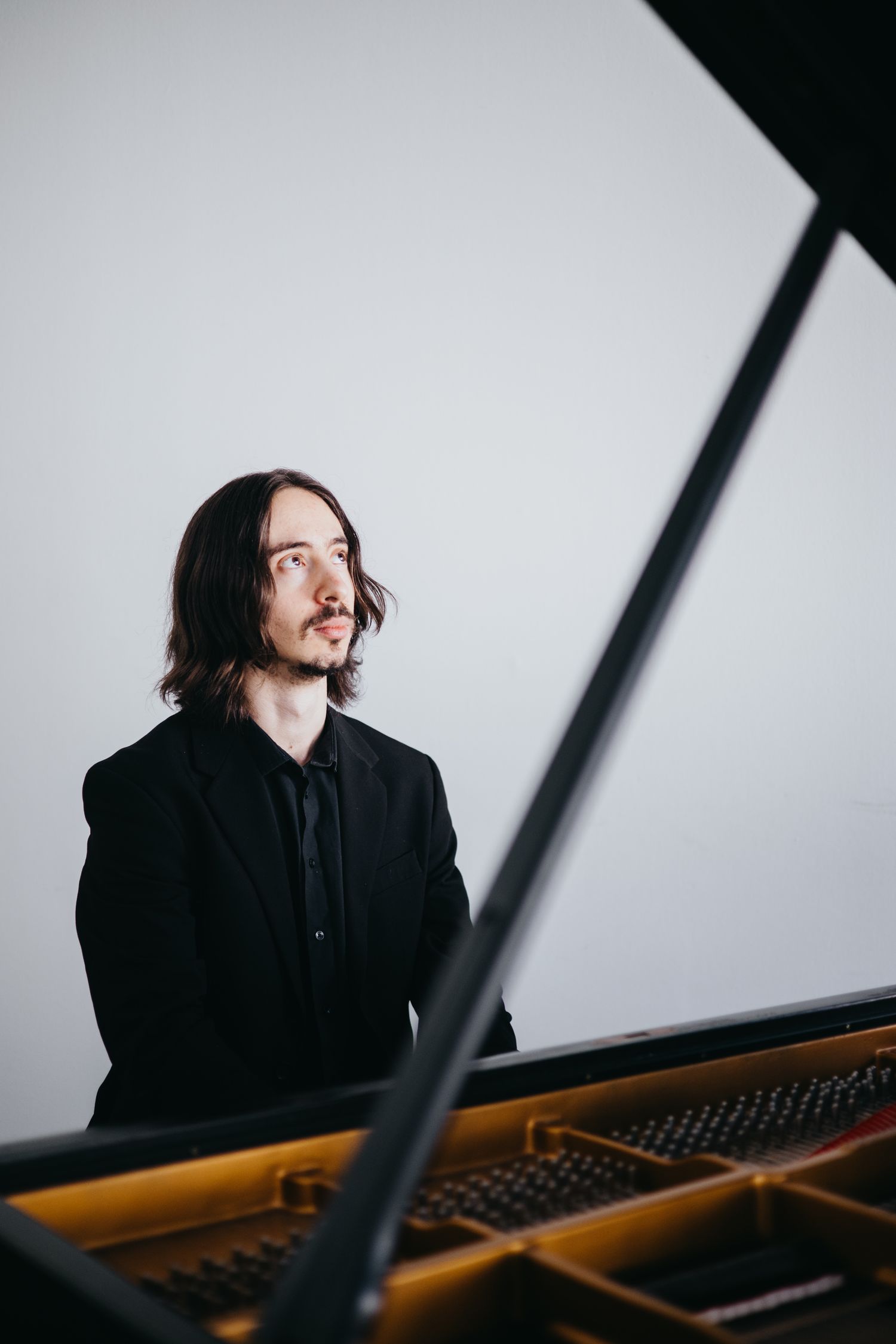 Male pianist in black suit sitting pensively beside an open grand piano in a studio.