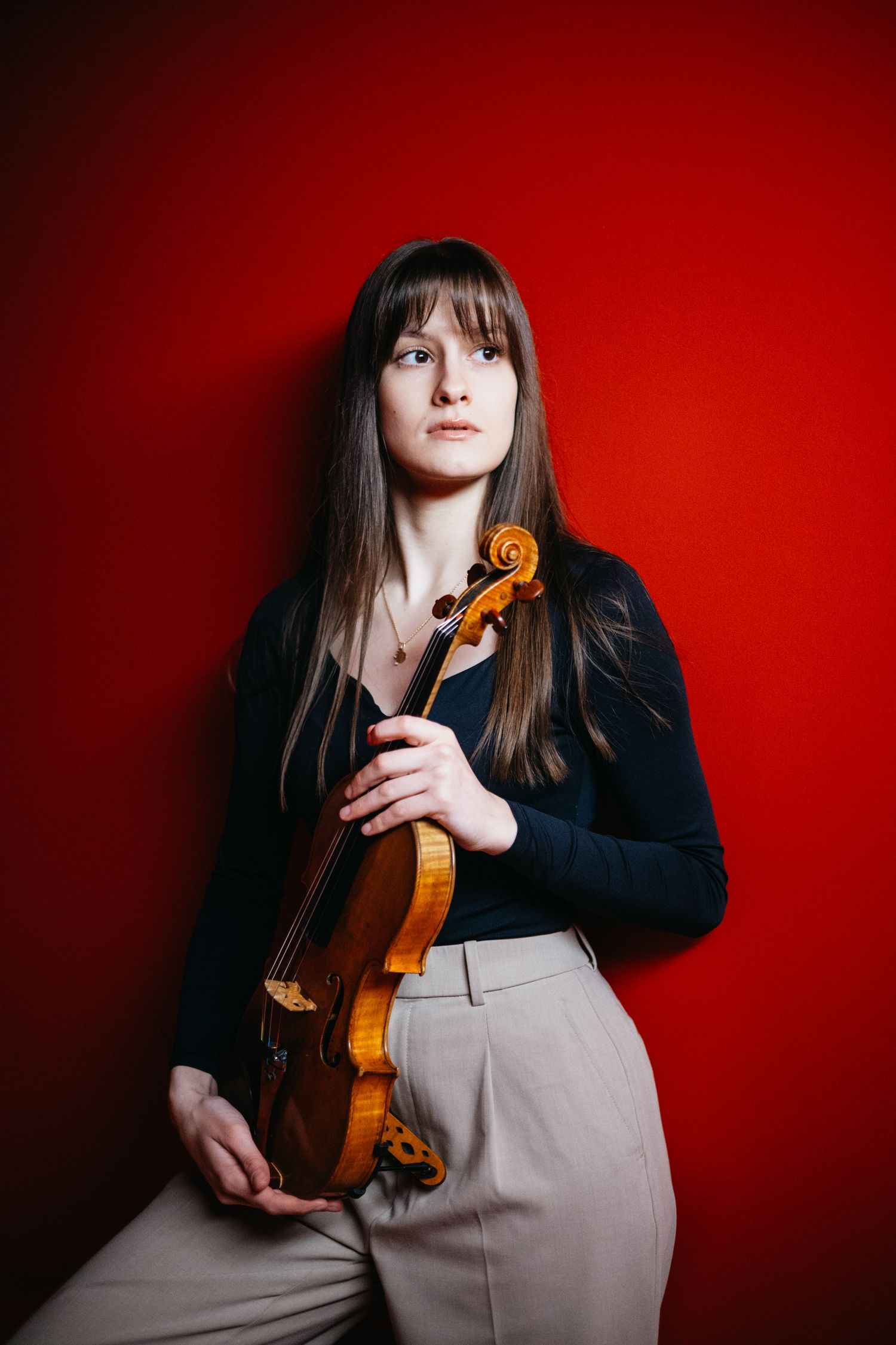 Female violinist holding a violin against a bold red background in a black top.