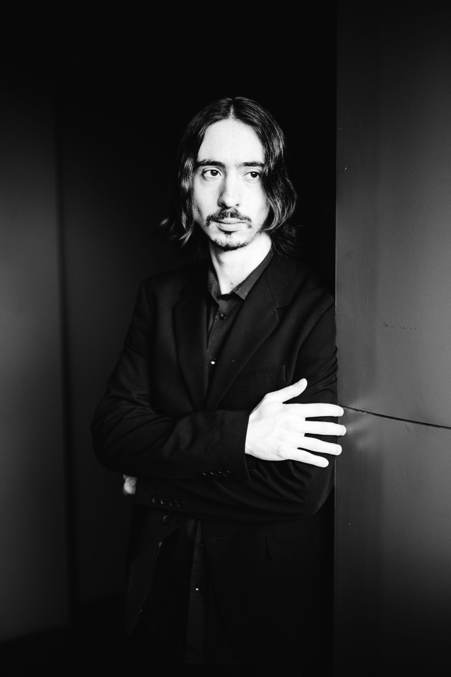 Black and white portrait of a long-haired male pianist in black suit with arms crossed.