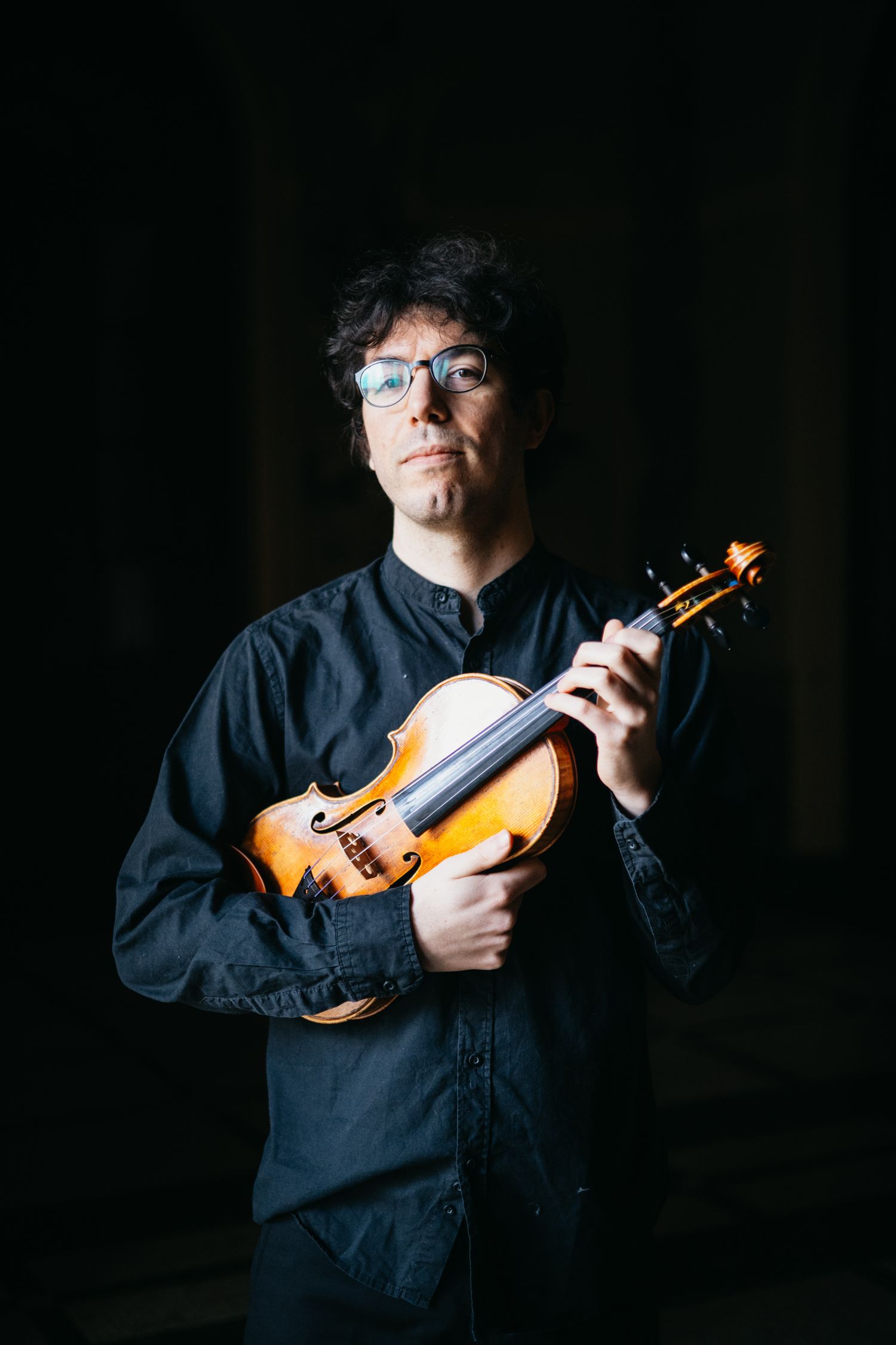 Violinist in black shirt and glasses holds violin against dark background.