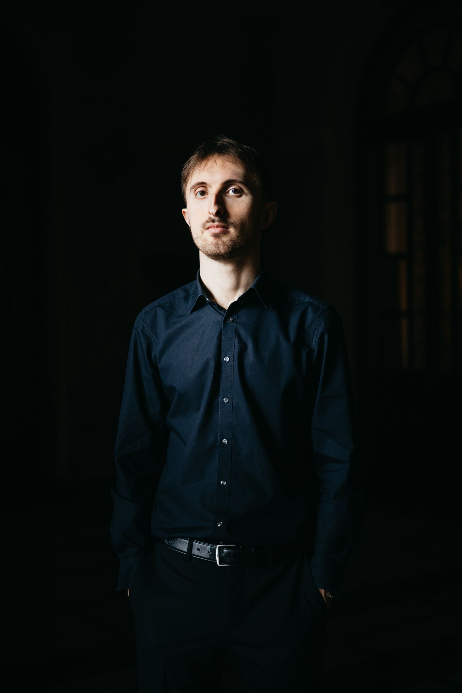 Young man in navy blue shirt standing confidently in dramatic low-key dark studio portrait.
