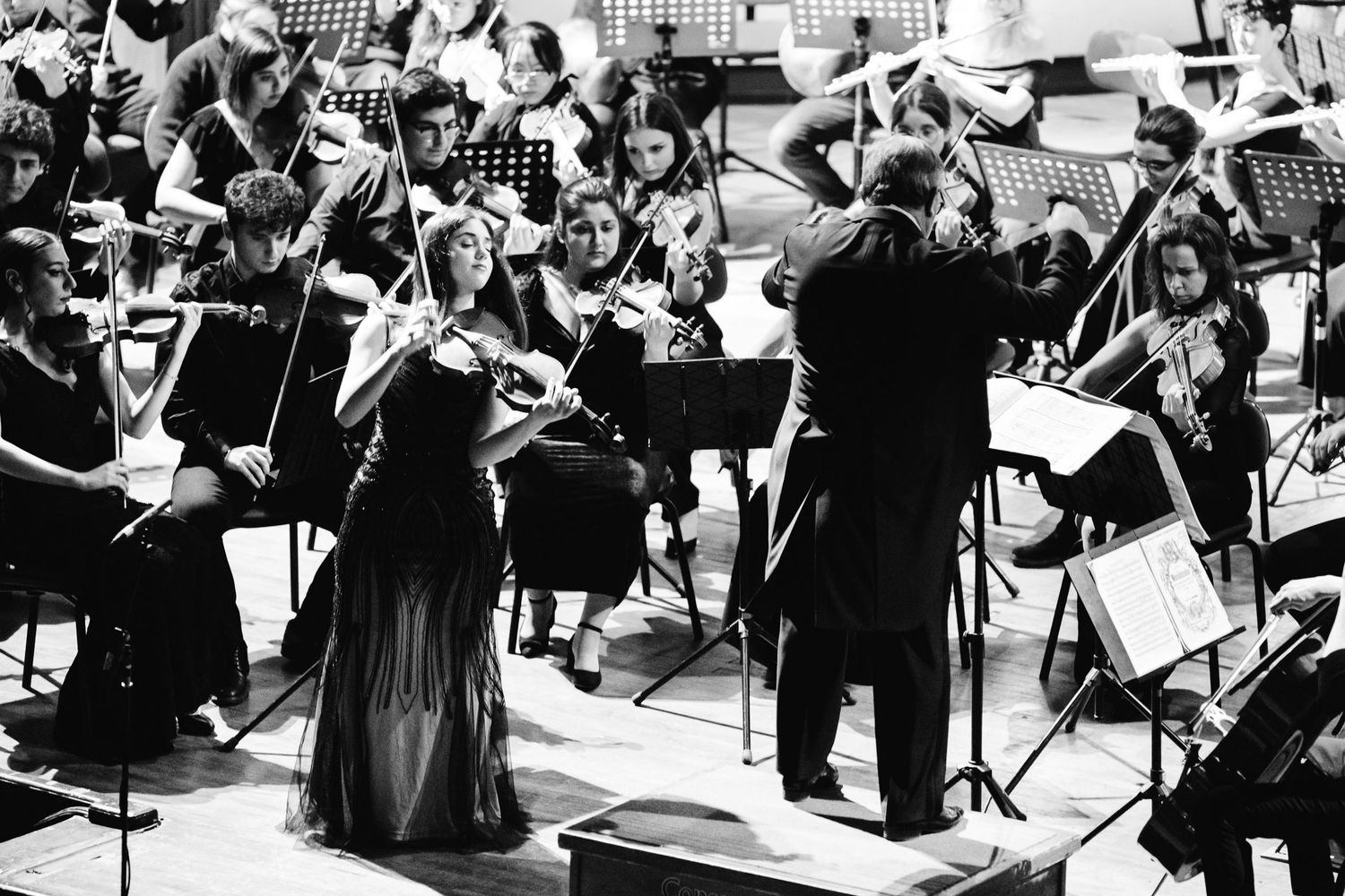 A female violinist performs as a soloist with a full orchestra during a black and white concert performance.
