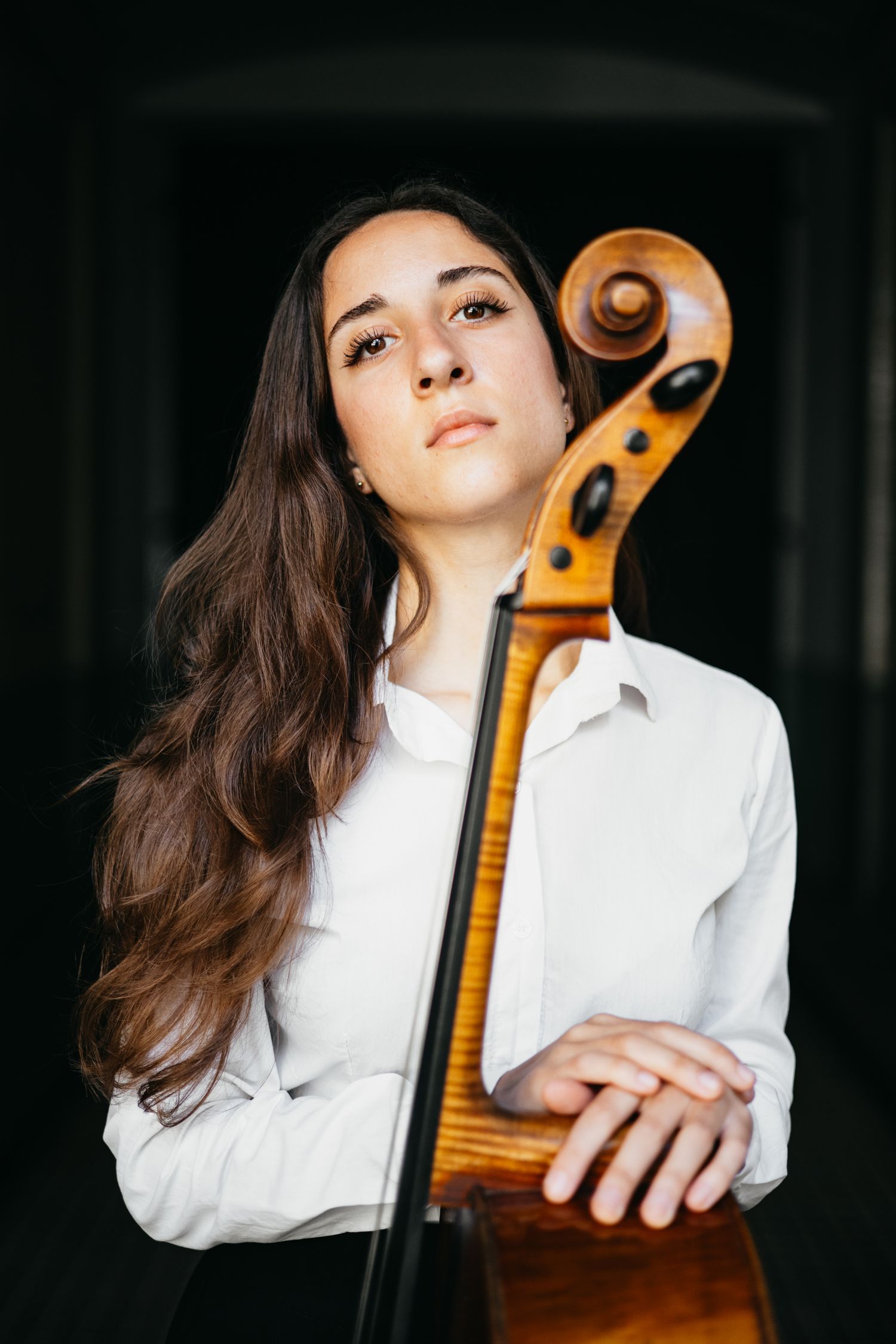 Portrait of a young female cellist in white shirt holding her cello in soft lighting.