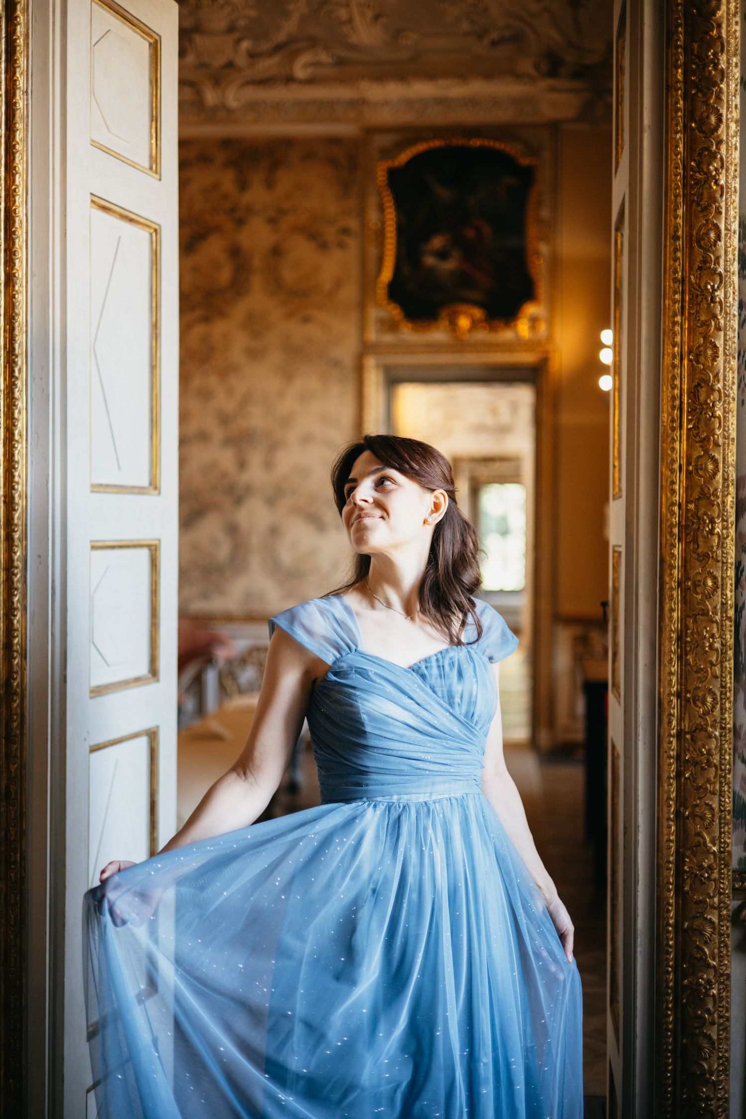 Woman in blue tulle gown poses gracefully in ornate doorway of elegant vintage room.
