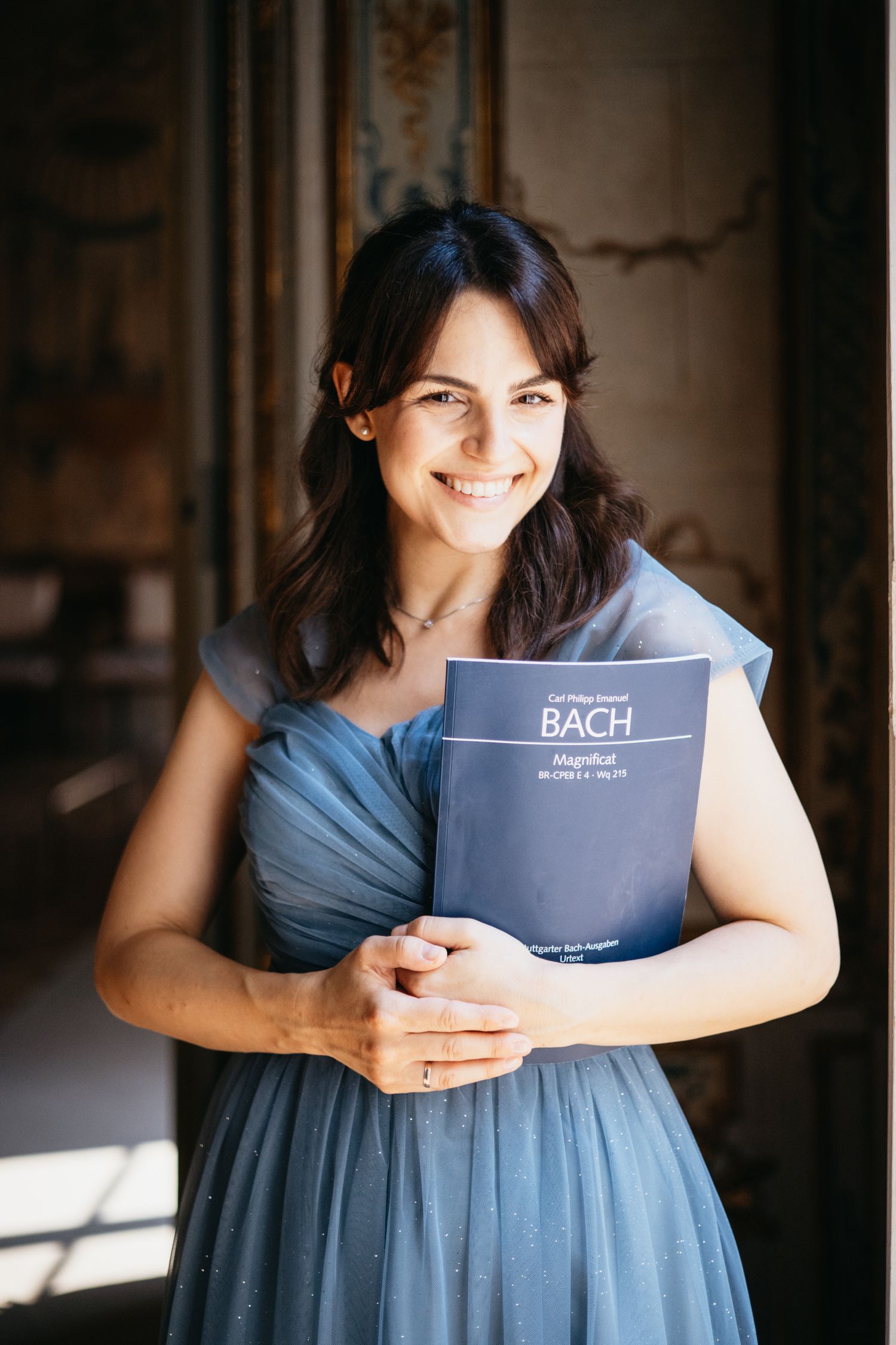 Smiling woman in blue dress holding a Bach Magnificat music score sheet, standing in an elegant interior.