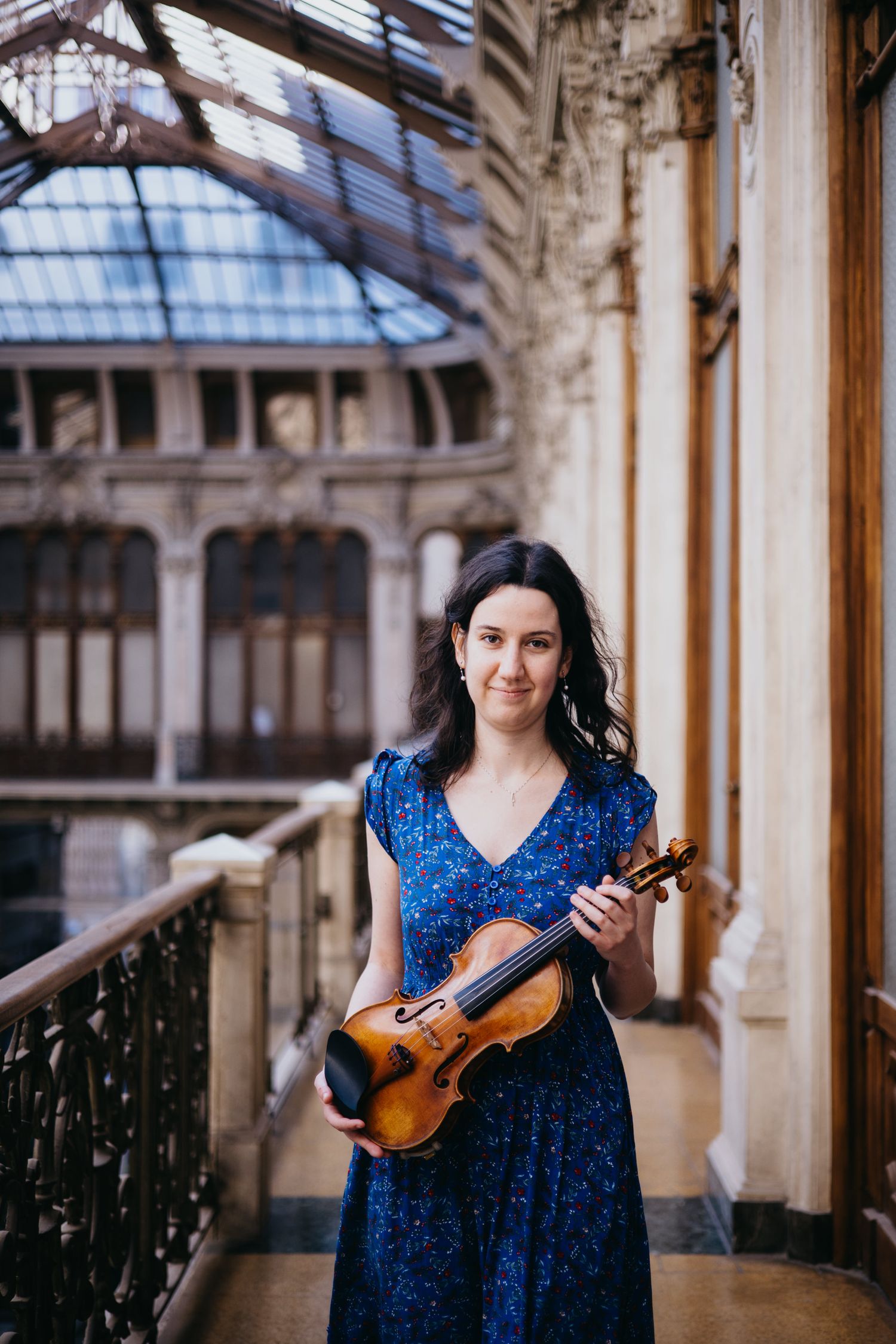 Woman in blue dress holding a violin in an elegant historic building with glass ceiling.
