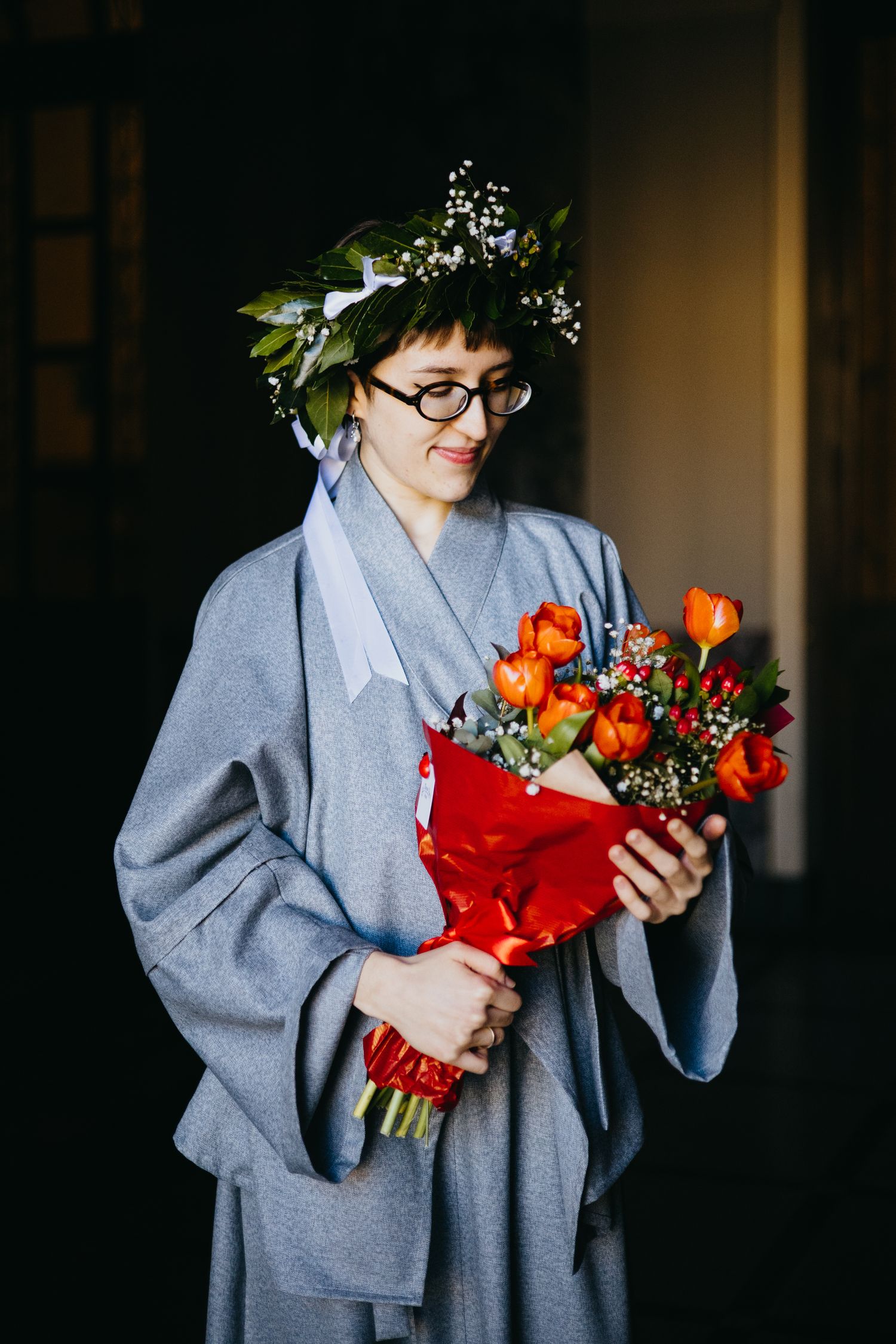 Graduate in gray robe with floral crown gazes at colorful orange tulip bouquet indoors.