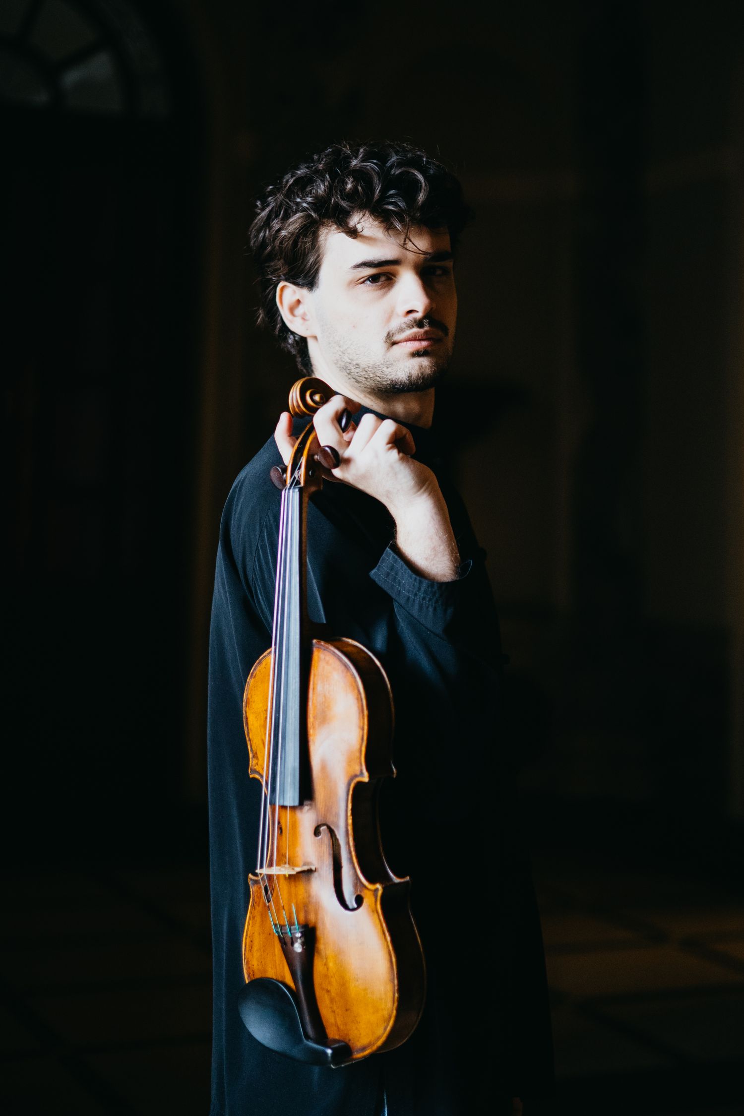 Serious male musician in black shirt poses with violin against dark background.