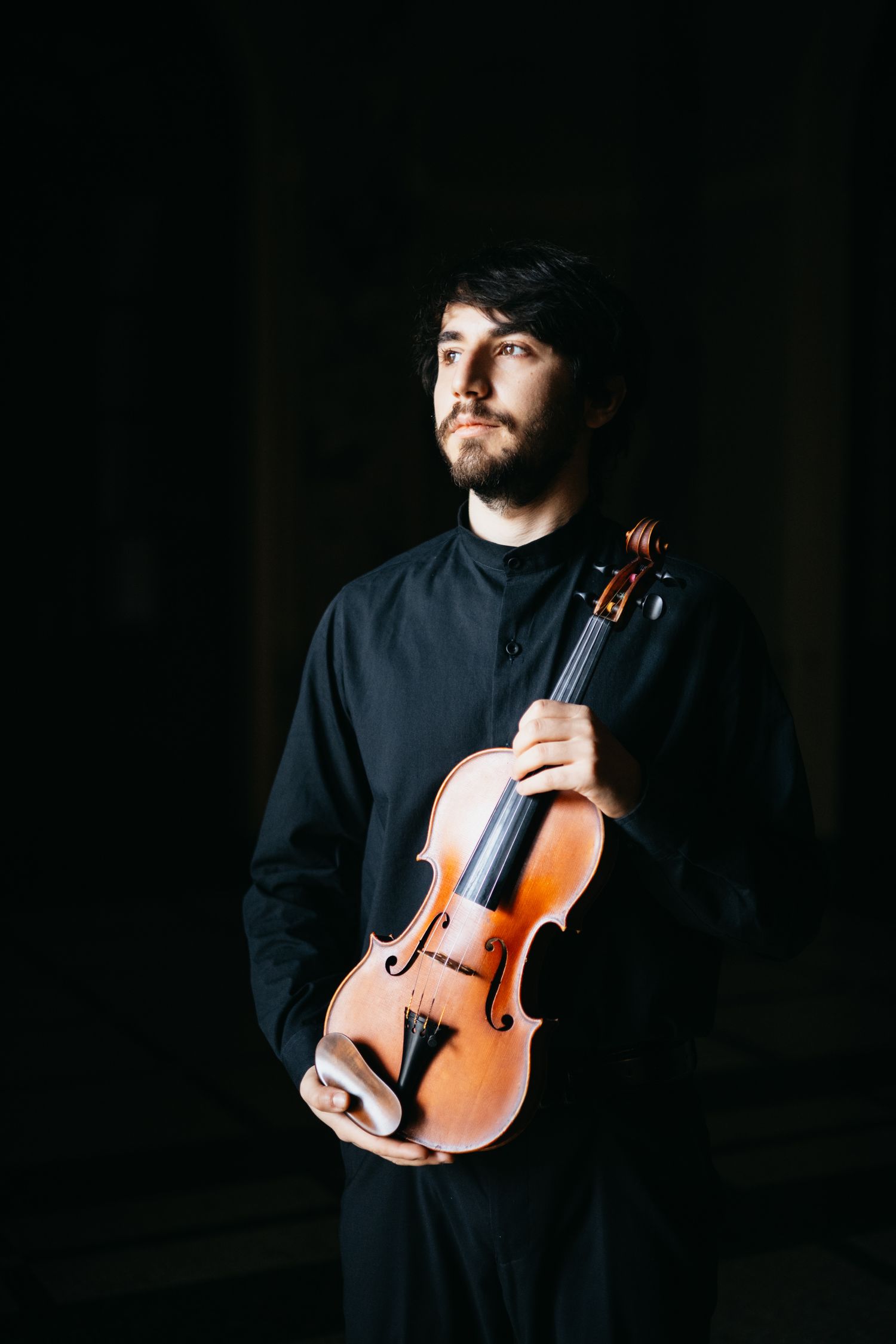 Male musician in black shirt holding violin against dark background in moody portrait.