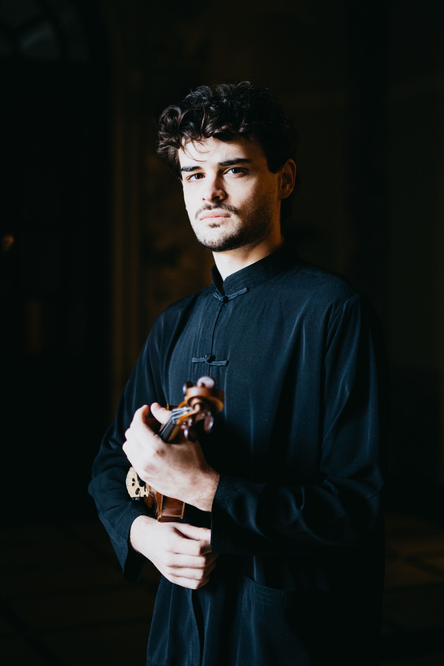 Young male violinist in black mandarin collar shirt holds violin in dramatic lighting.