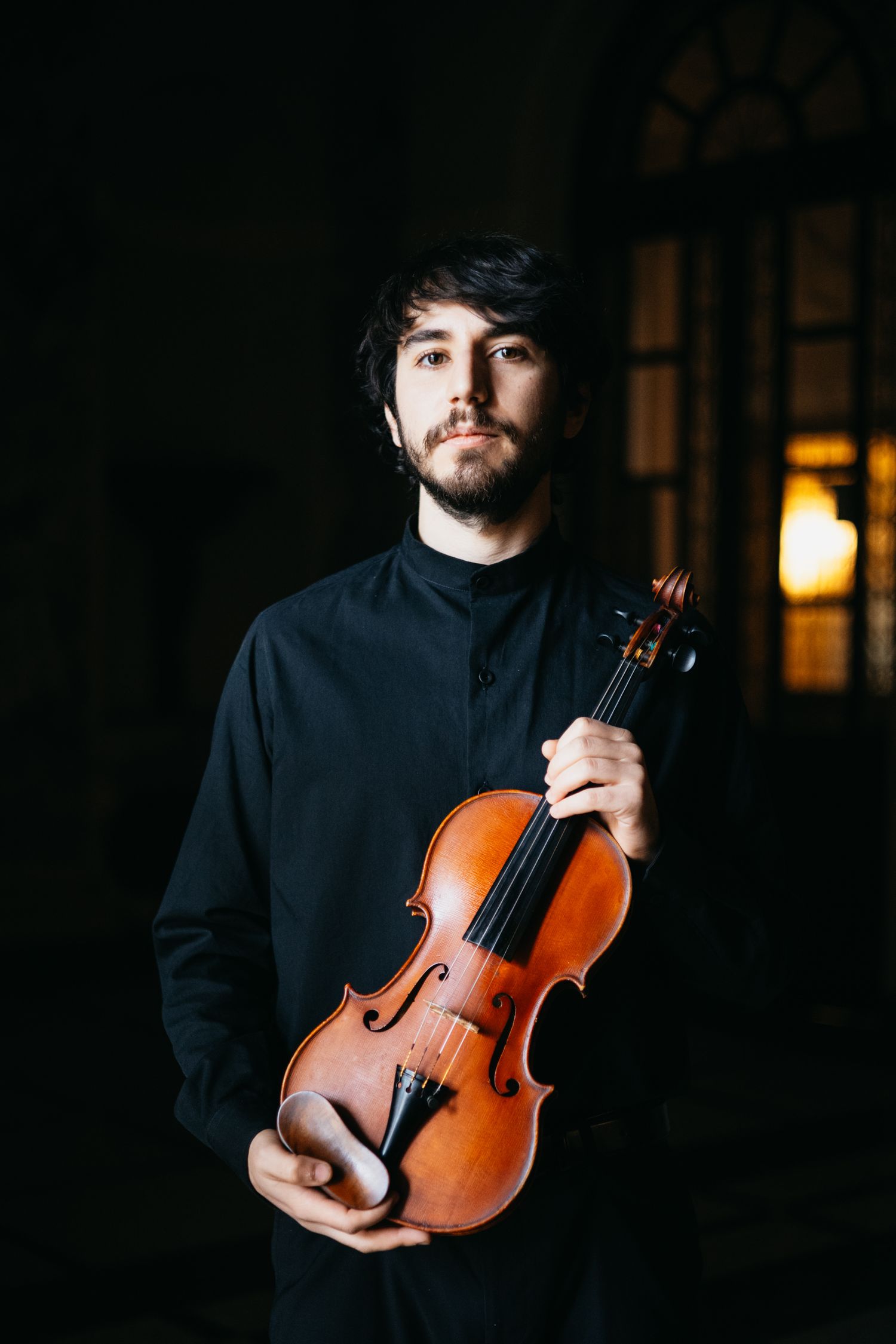 Male violinist in black outfit holding violin while looking at camera in dimly lit setting.