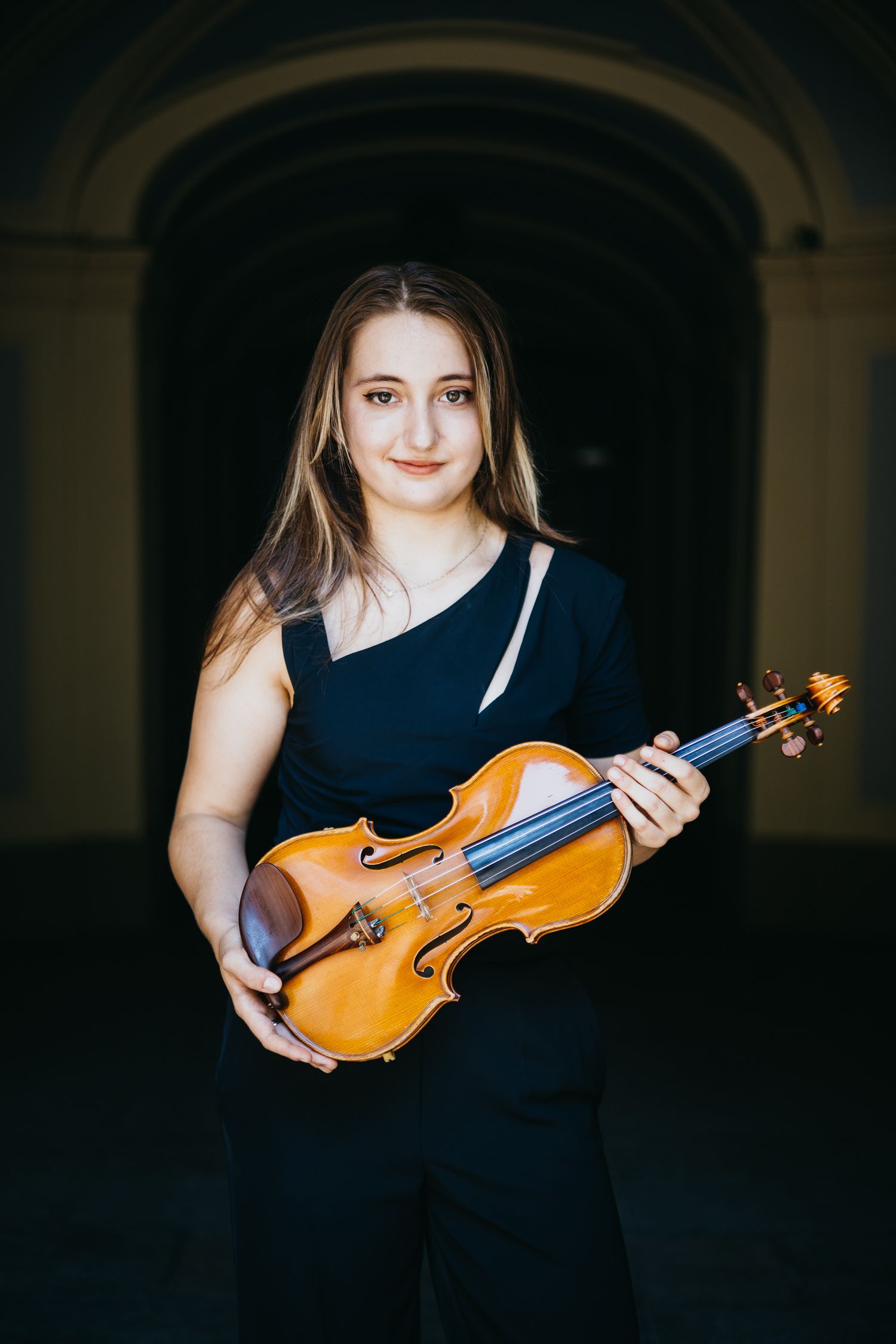 Smiling young woman in black outfit holding a violin in an arched corridor.