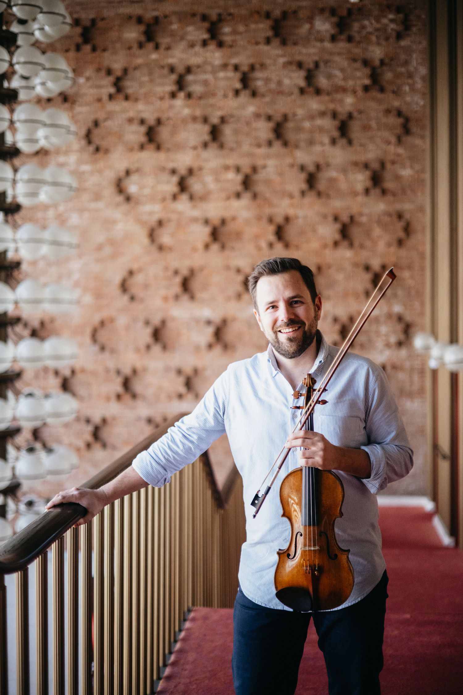 Smiling male violinist holding violin and bow in white shirt, posing in elegant venue with brick wall backdrop.