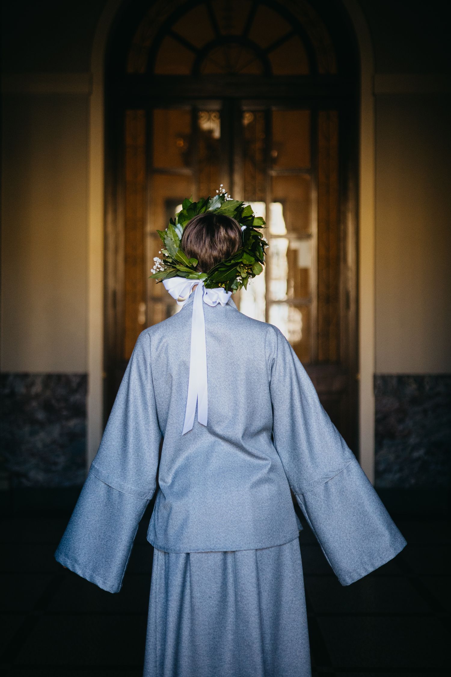 Back view of woman in gray coat with floral crown and white ribbon bow.