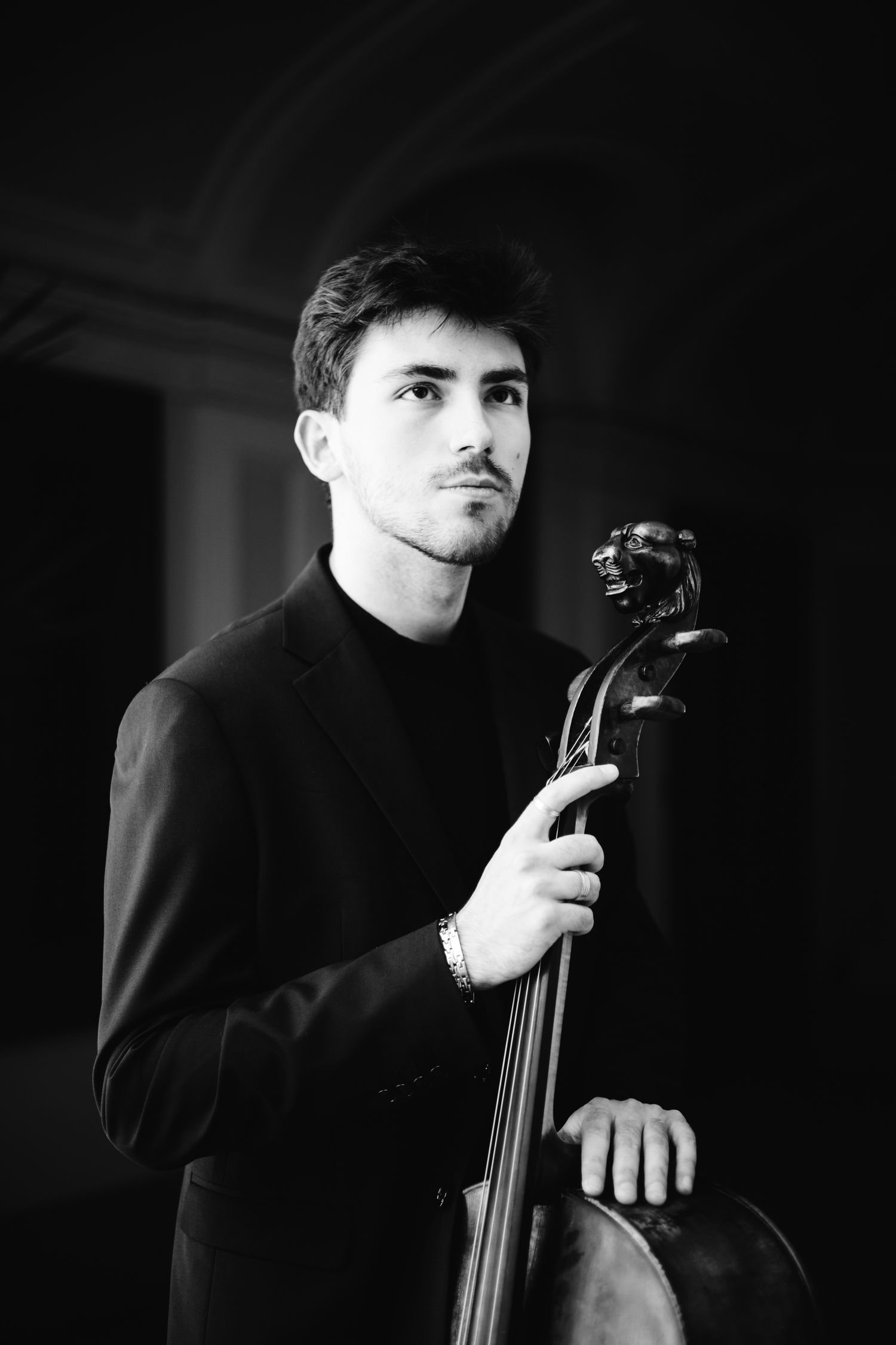 Black and white portrait of male cellist holding cello scroll in dramatic low-key lighting.