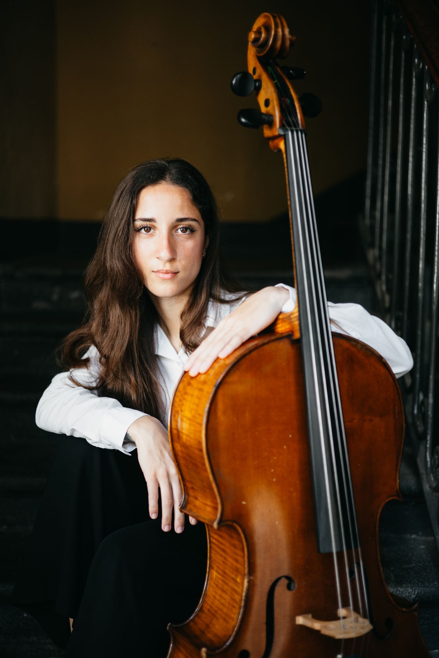 Young female cellist sitting on stairs, leaning against her cello in a vintage interior.