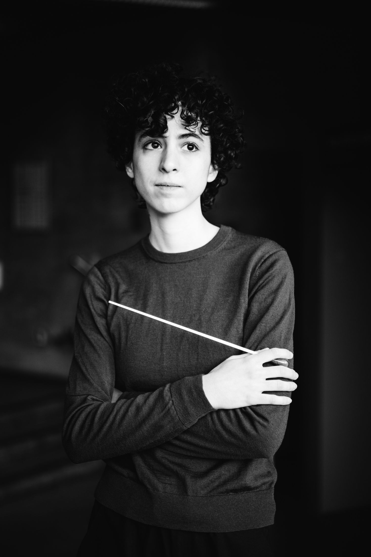 Black and white portrait of young conductor holding baton with arms crossed.
