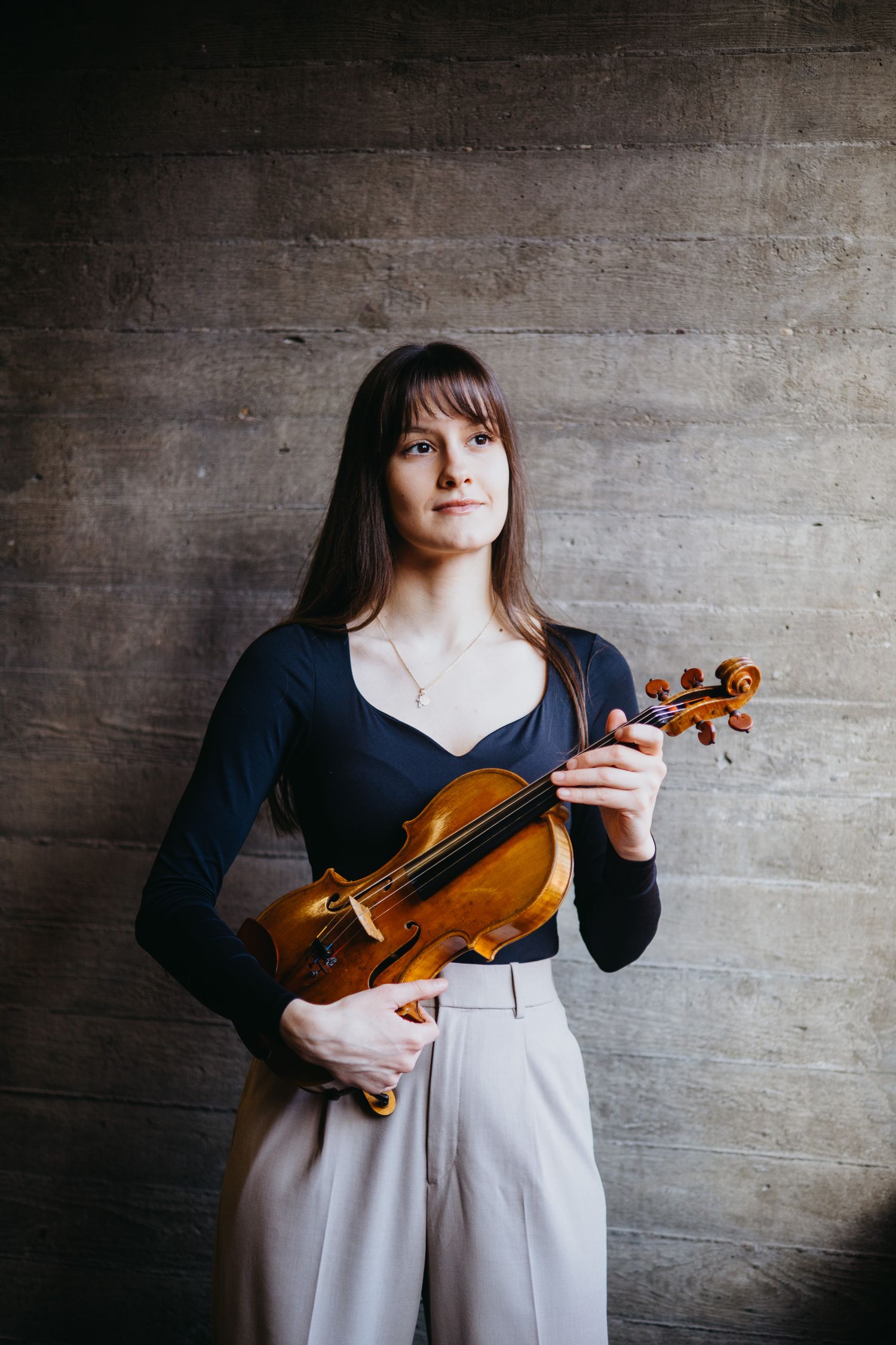 Female violinist in black top holding a violin against a textured concrete wall.