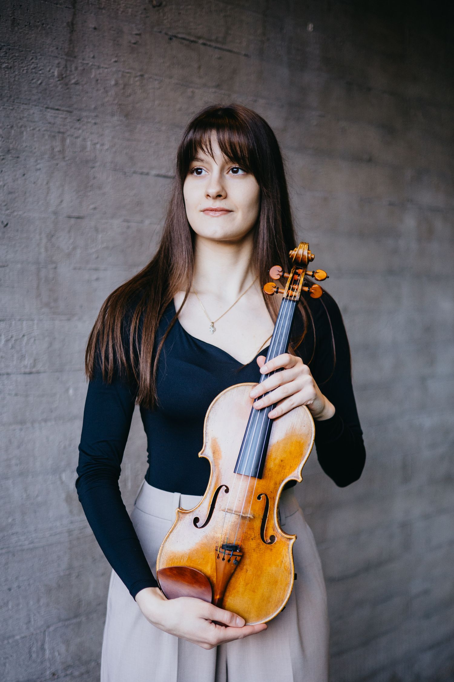 Young female violinist holding a violin against a textured concrete wall.