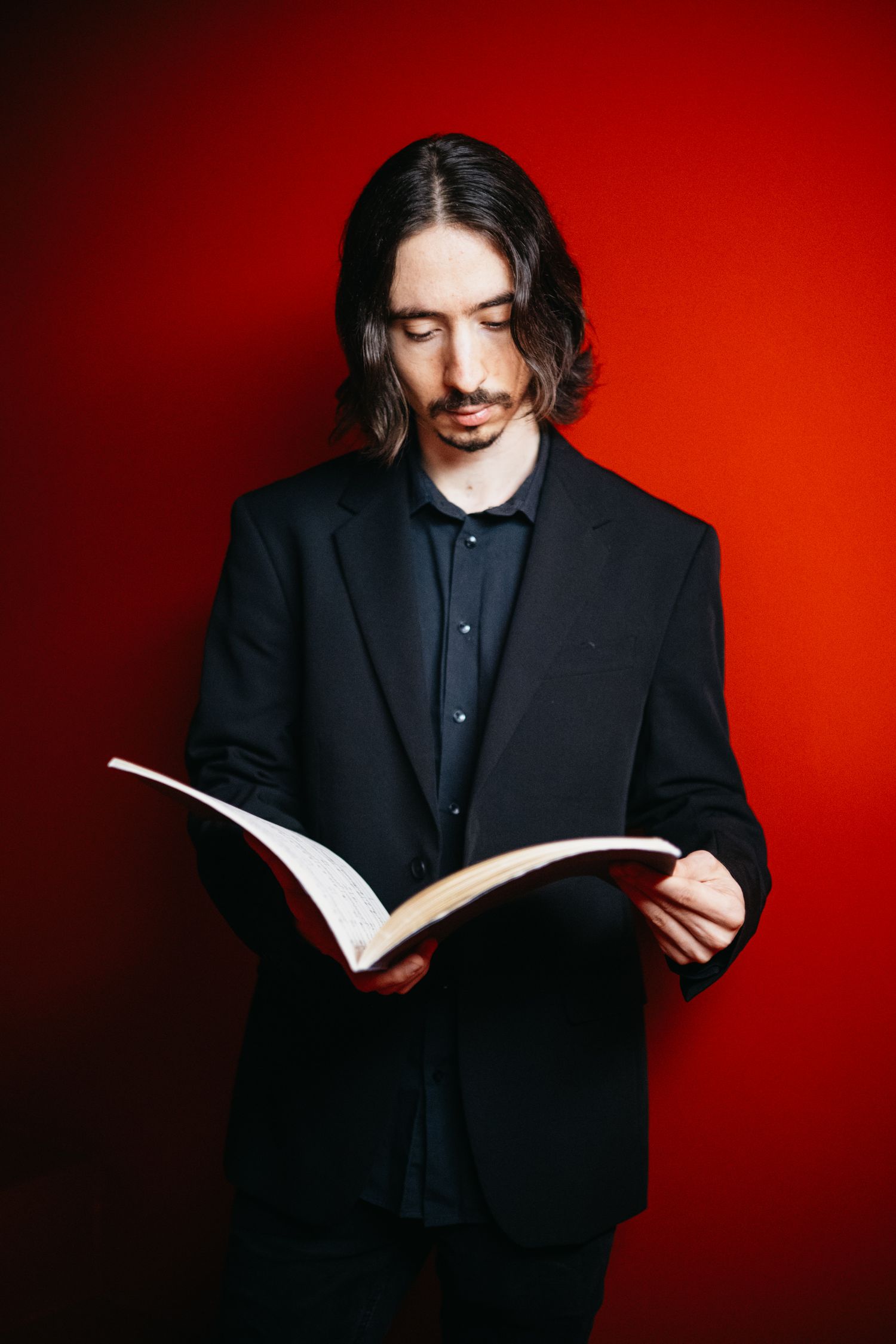 Long-haired male musician in black suit reading sheet music book on red background.