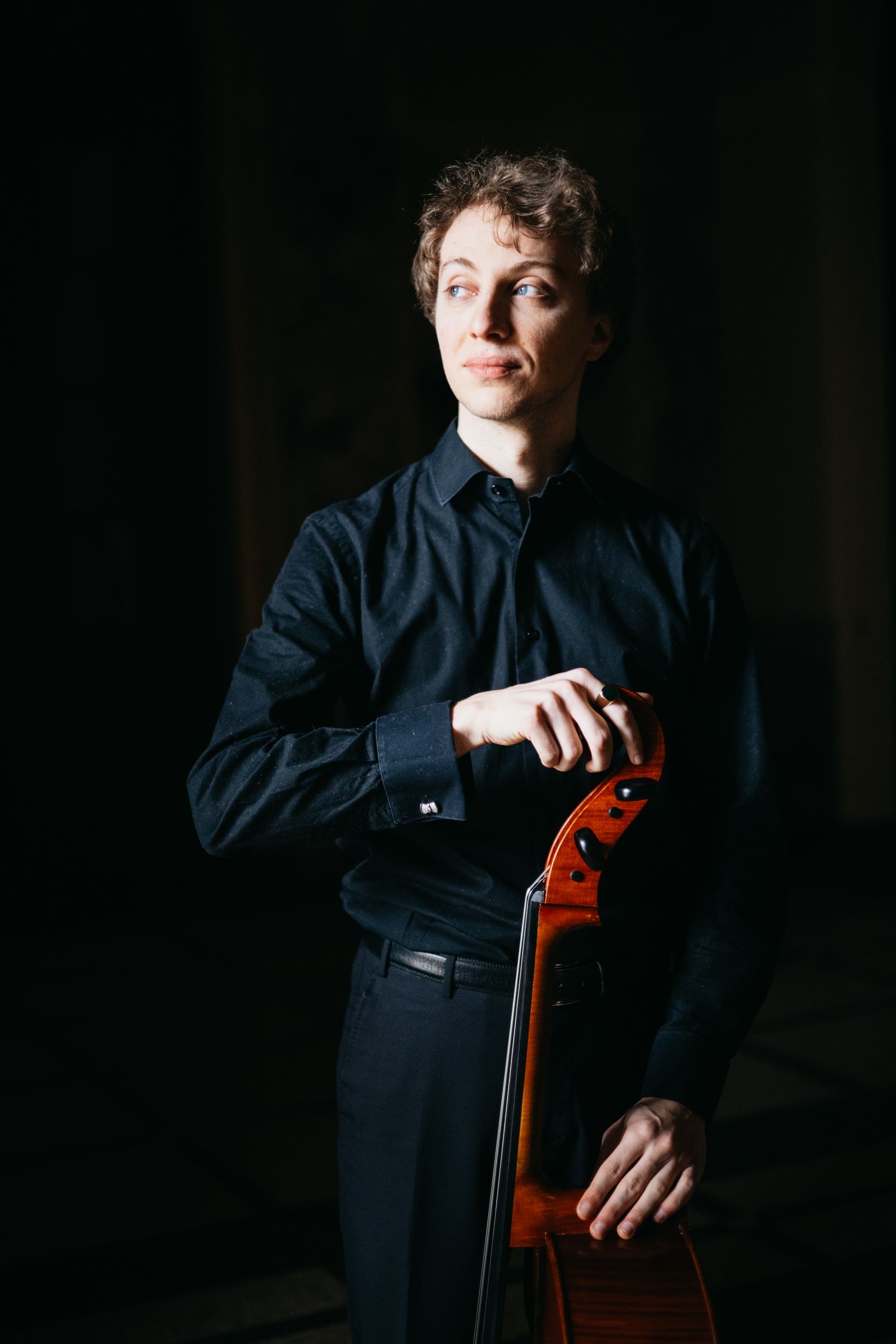 Male cellist leans on cello scroll wearing black shirt in dark studio.