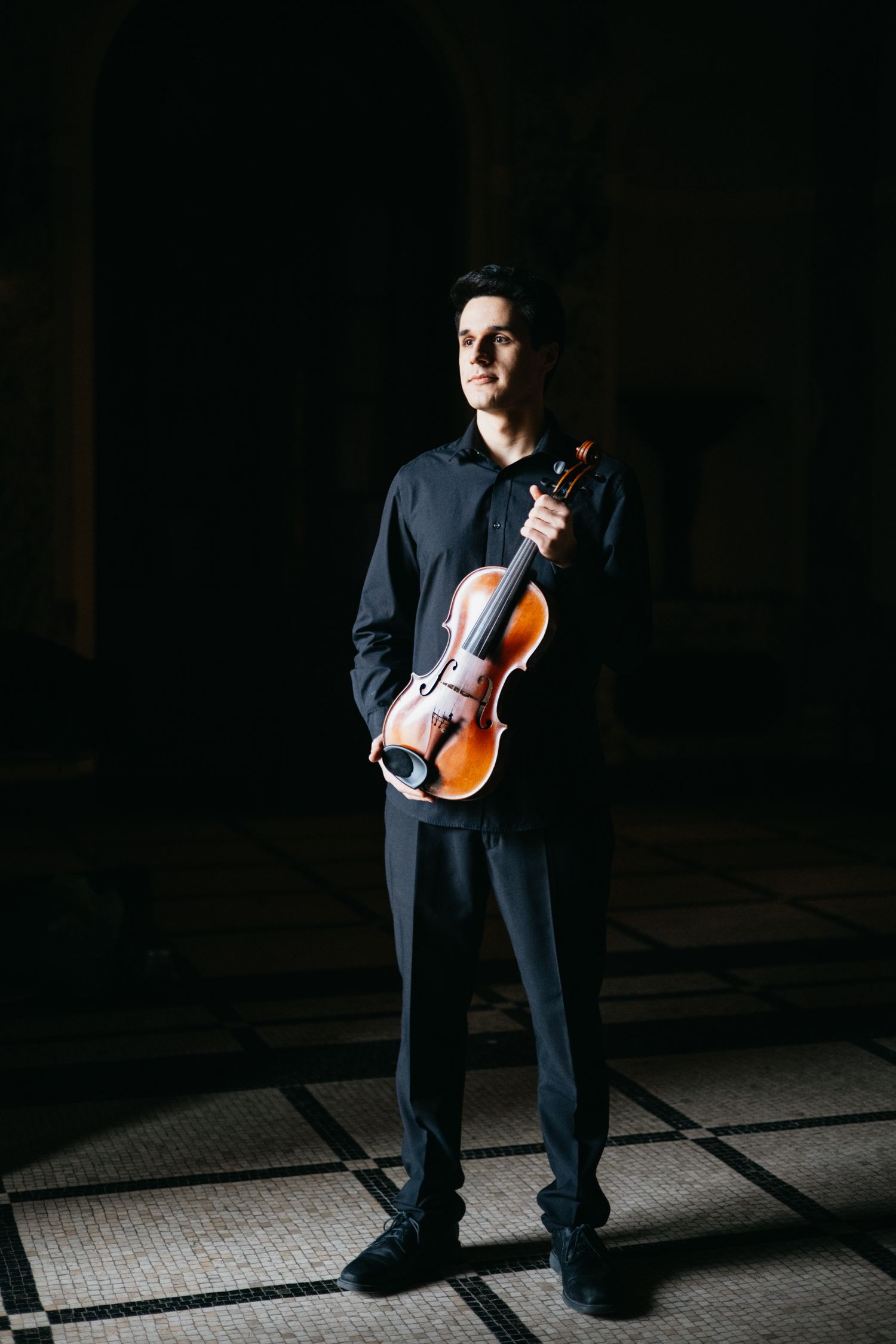 Full length portrait of male violinist in black holding violin in dark architectural space.