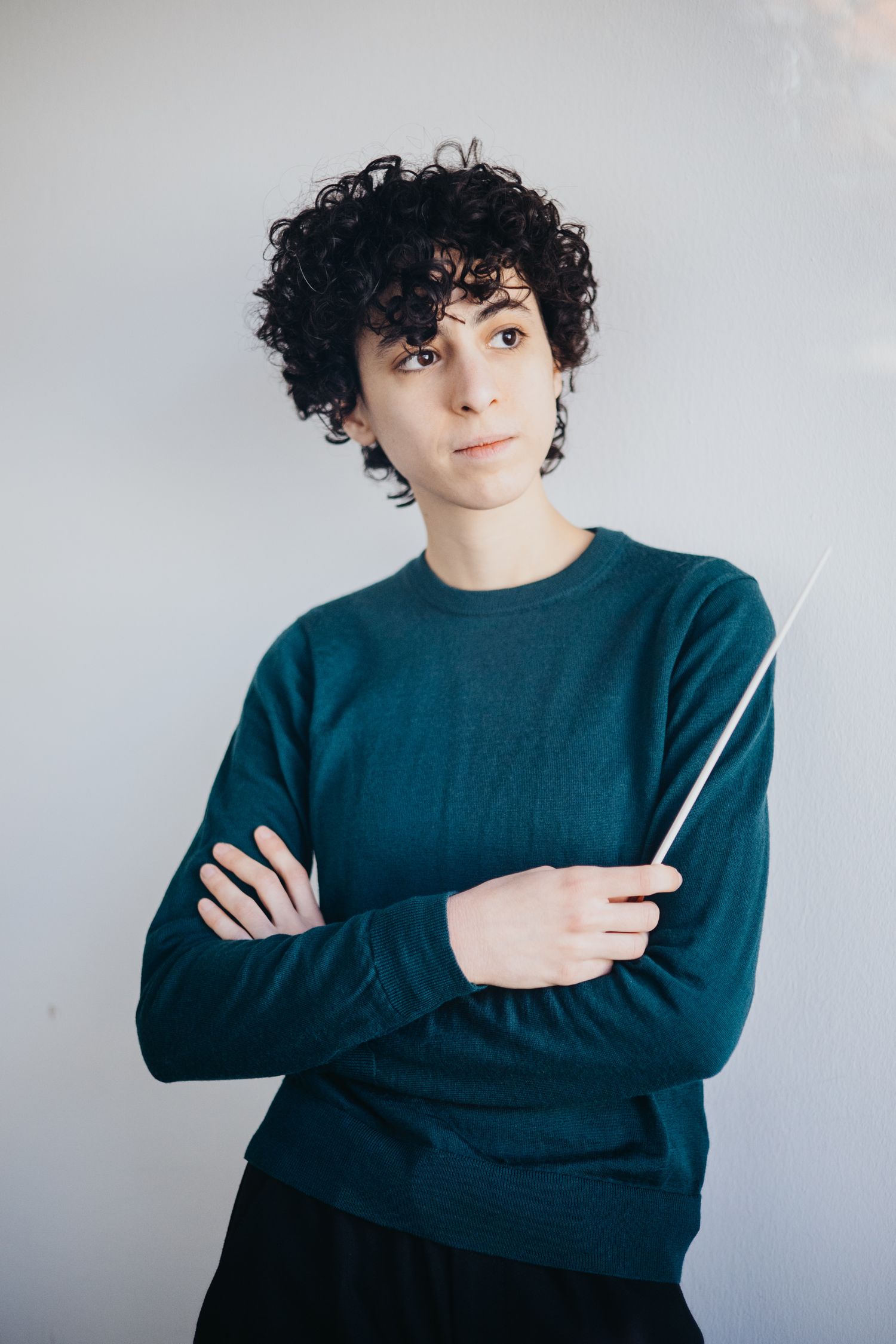Young conductor with curly hair holds a baton with arms crossed, wearing a teal sweater against a gray background.