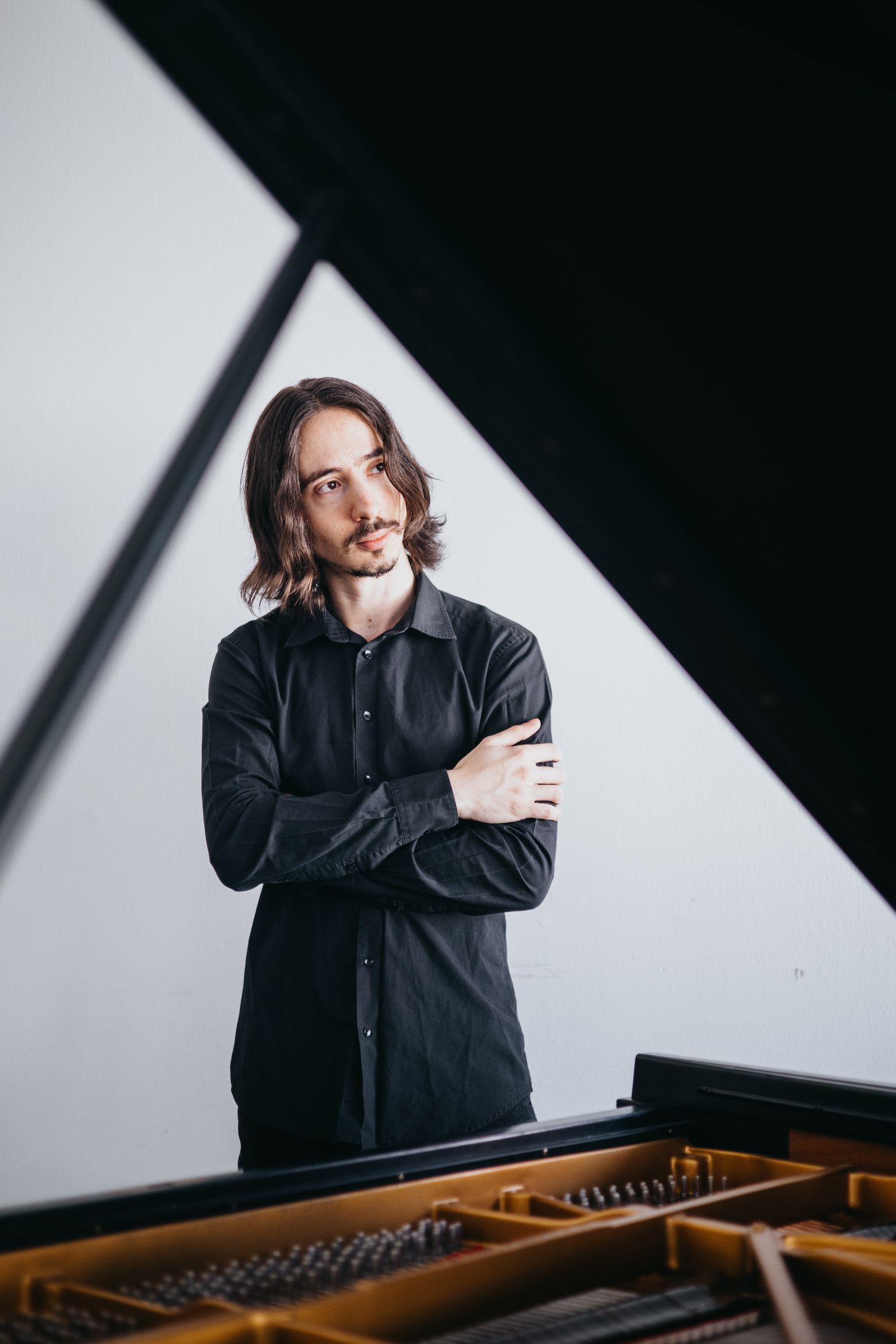 Long-haired male pianist in black shirt standing with arms crossed beside a grand piano.