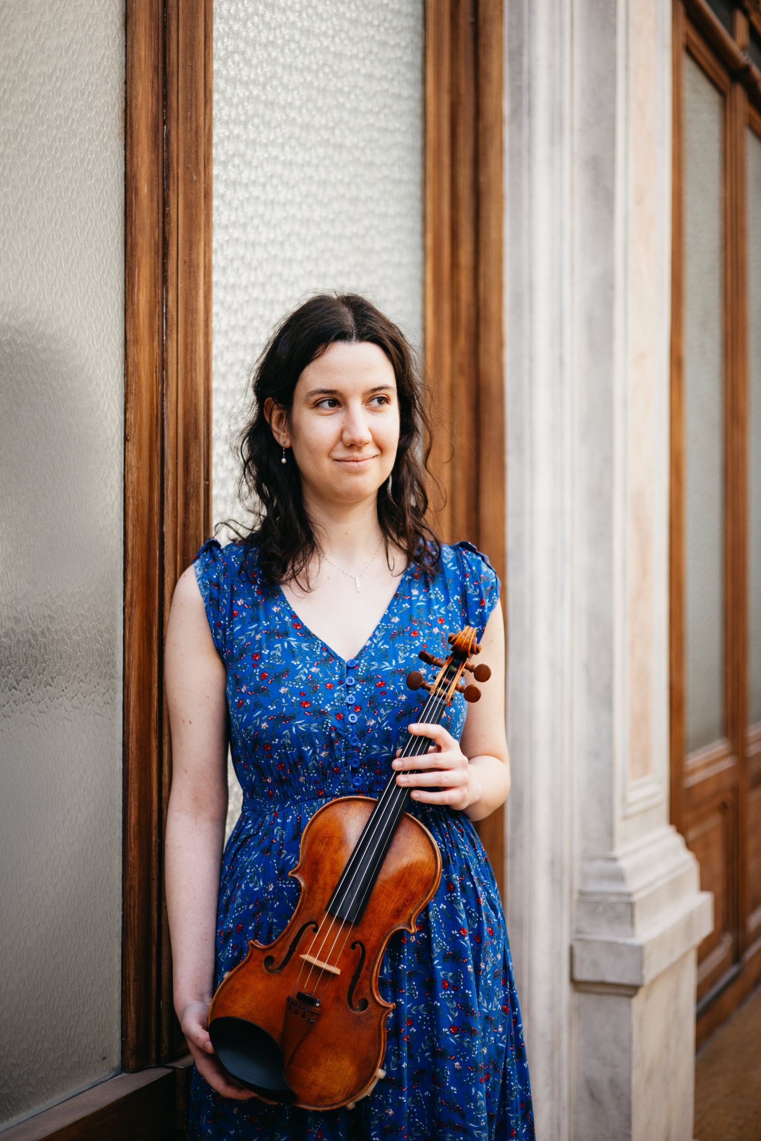 A young woman in a blue floral dress holds a violin while posing beside wooden columns and frosted glass.