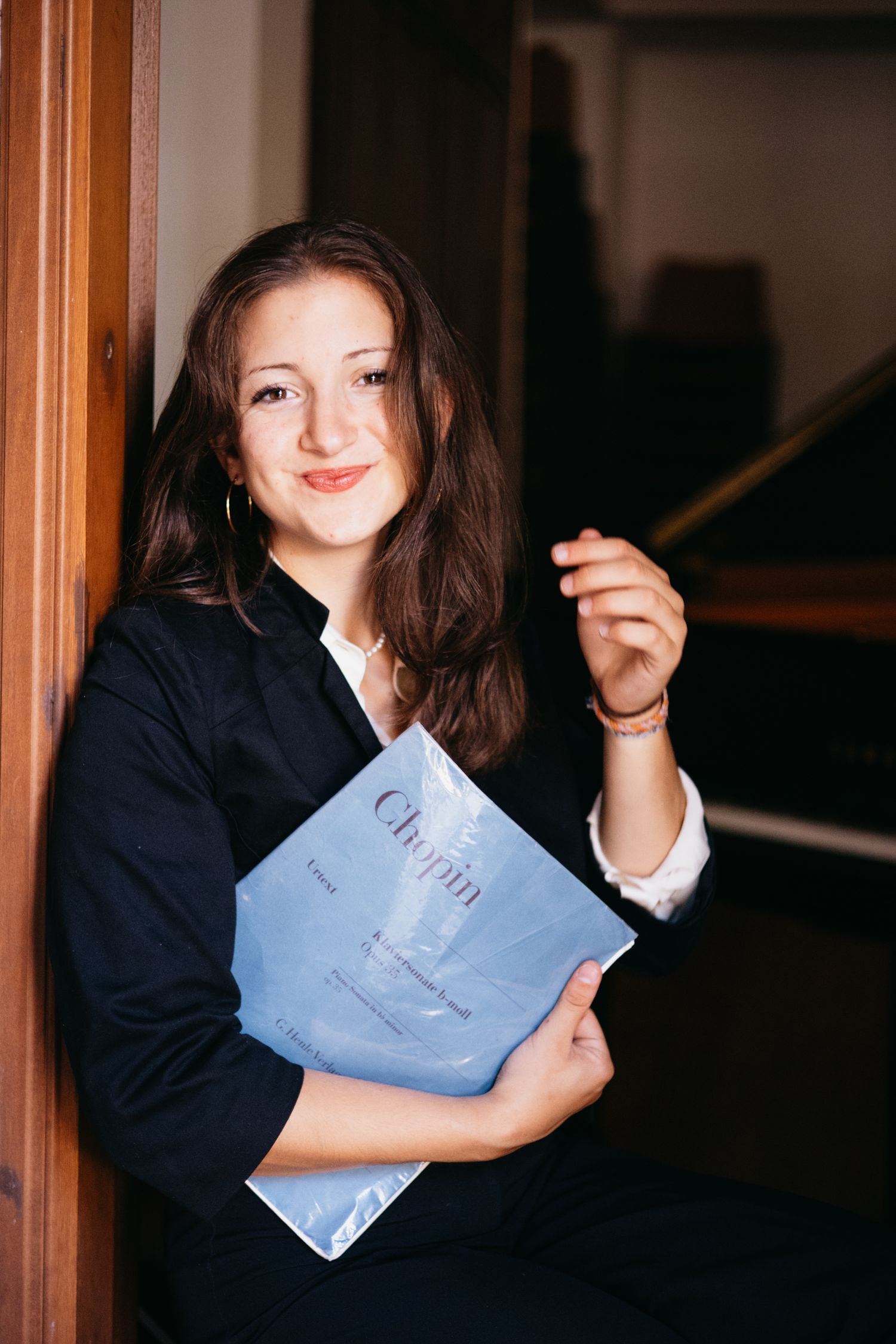Young woman smiling while holding a Chopin sheet music book, seated near a piano in a music room.