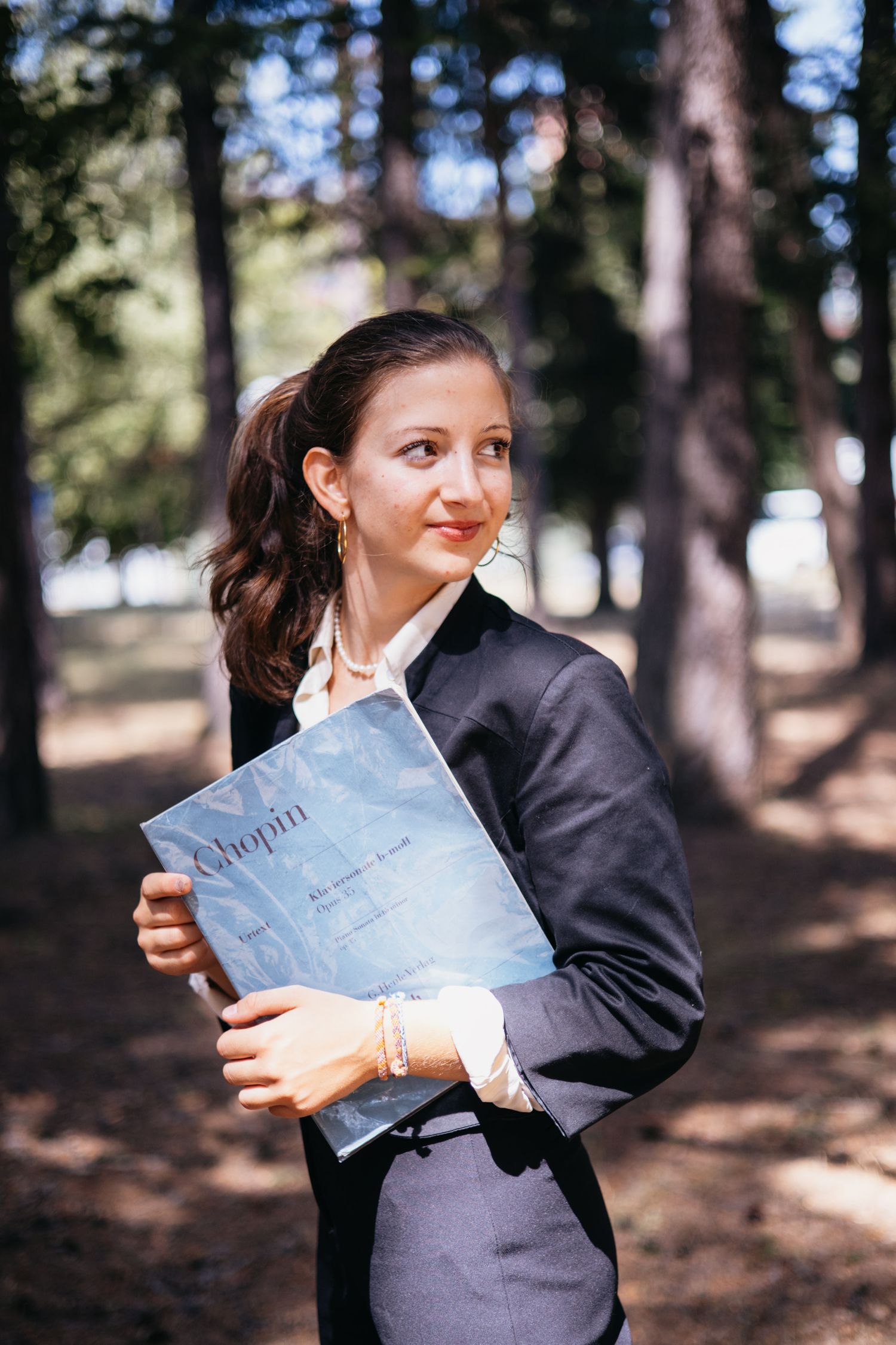 Young woman in blazer holding Chopin sheet music book, smiling outdoors in a sunlit pine forest.