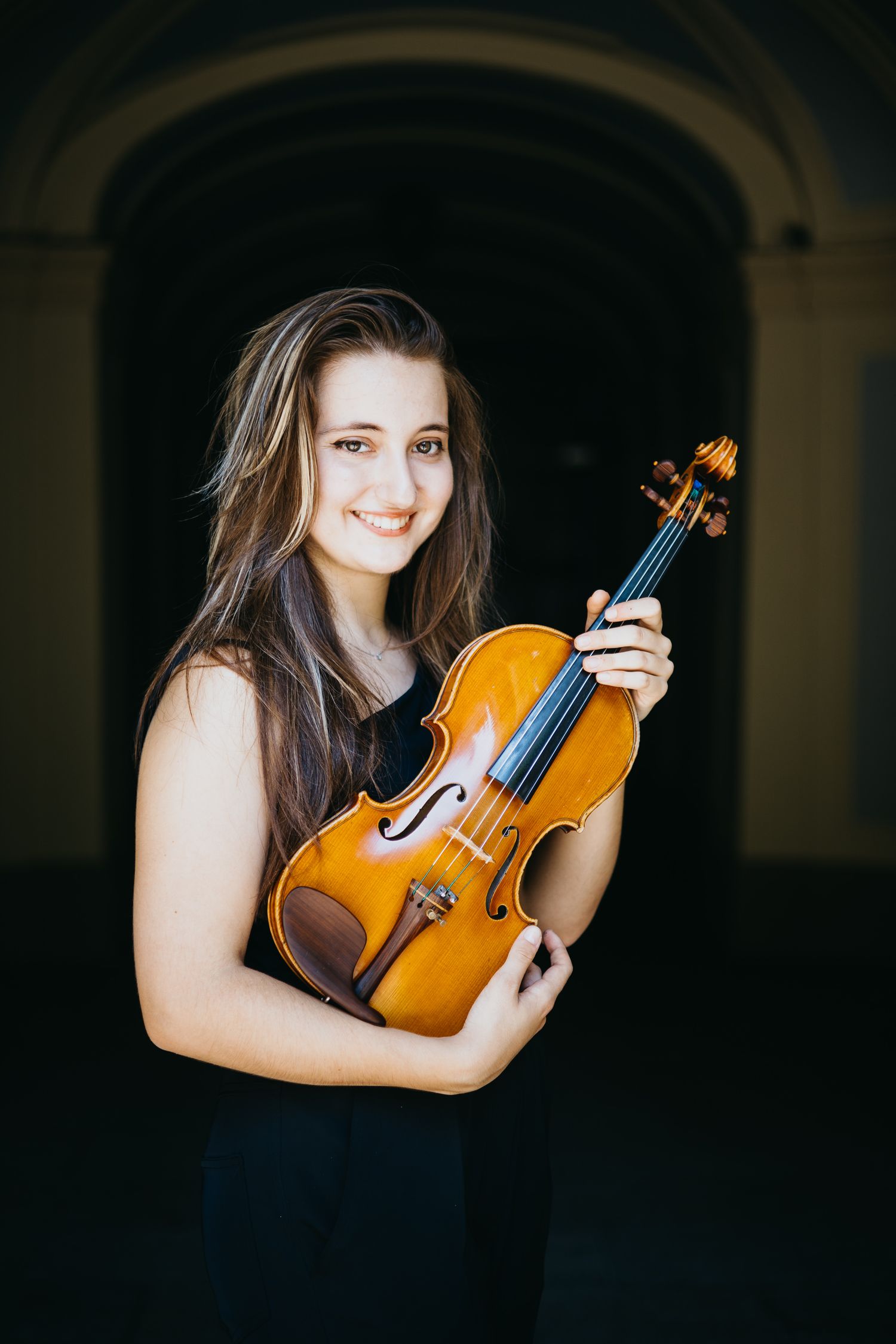 Smiling young female violinist in black dress holds violin under an arched doorway.