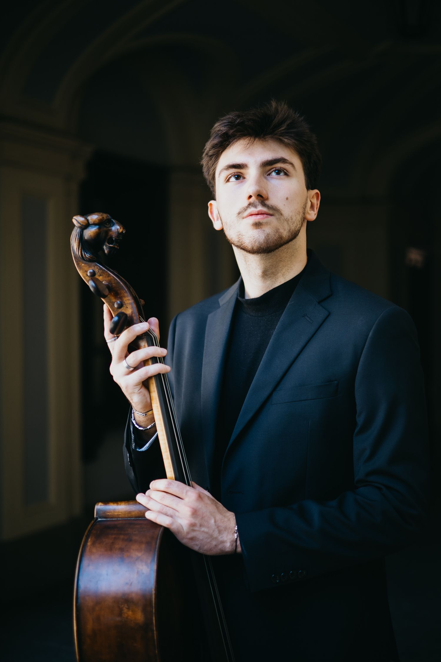 Young male musician in black suit holding cello scroll upright in dramatic arched interior.