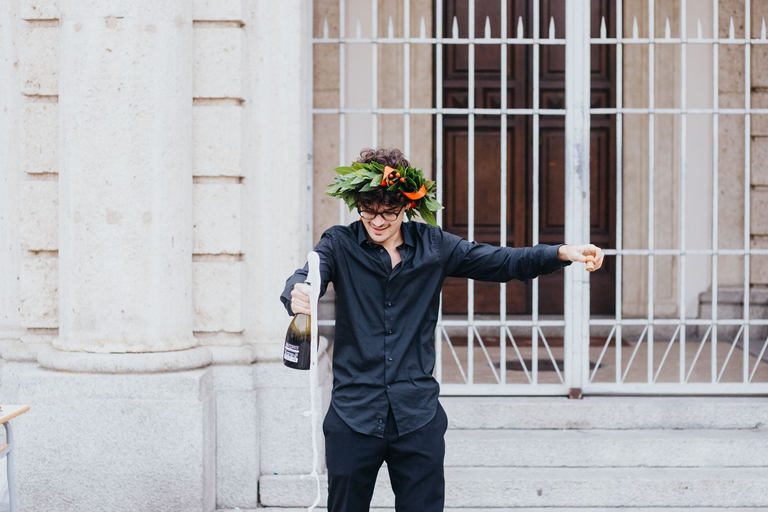 Graduate in laurel wreath poses playfully holding champagne bottle outside a classical building.