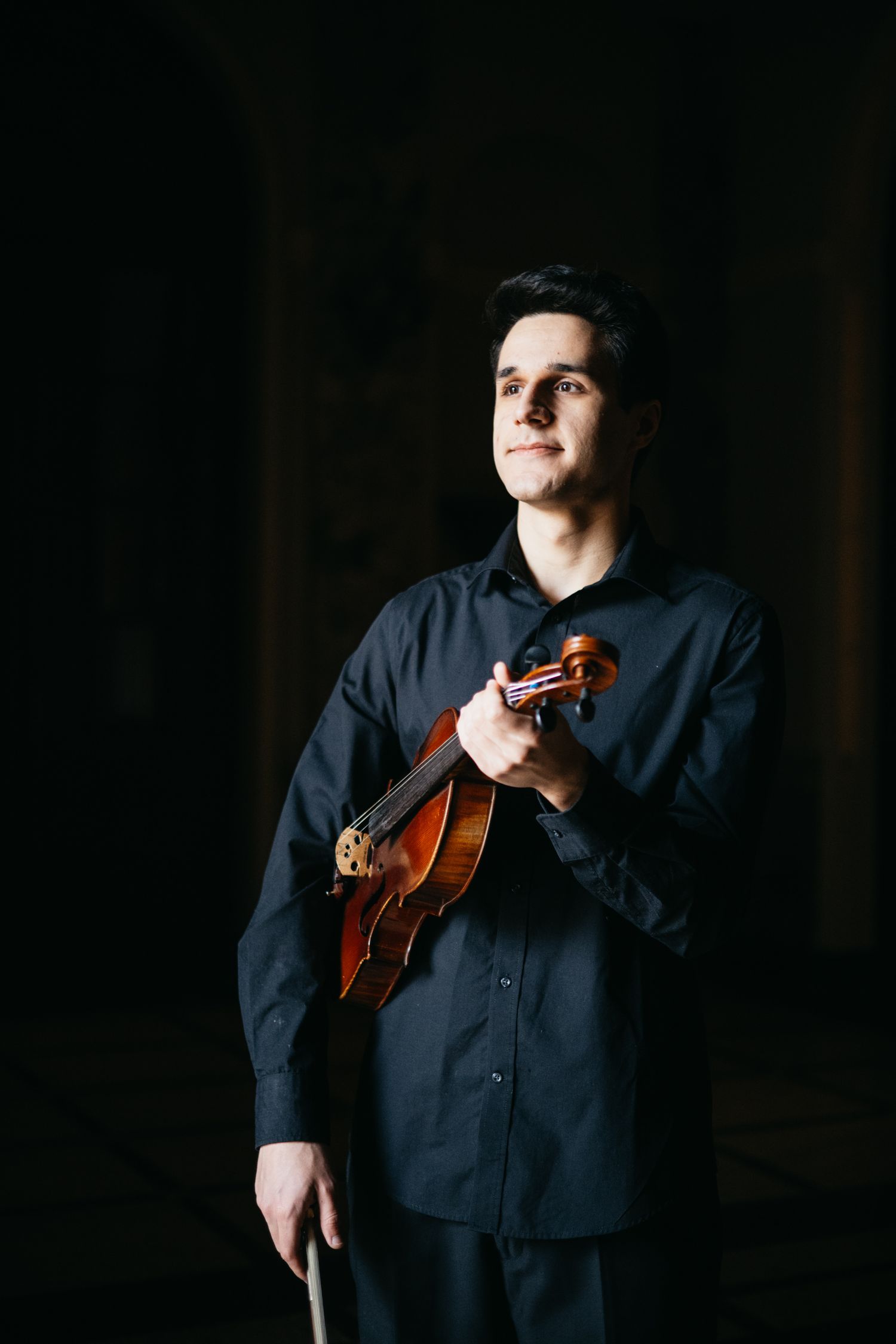 Young male violinist in black shirt holds violin against dark background in portrait.