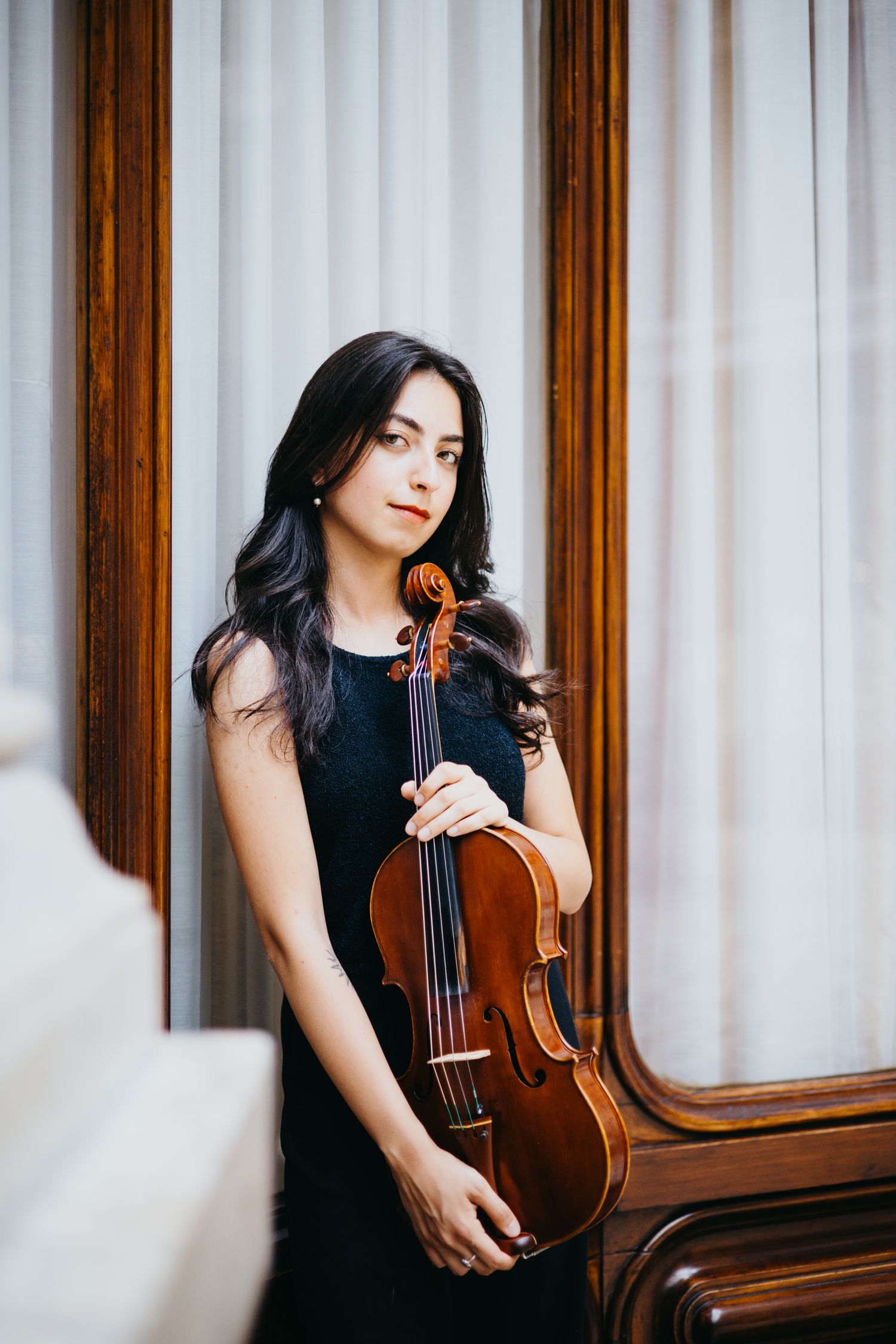 Female musician in black dress holds violin while standing by ornate wooden mirror frame.