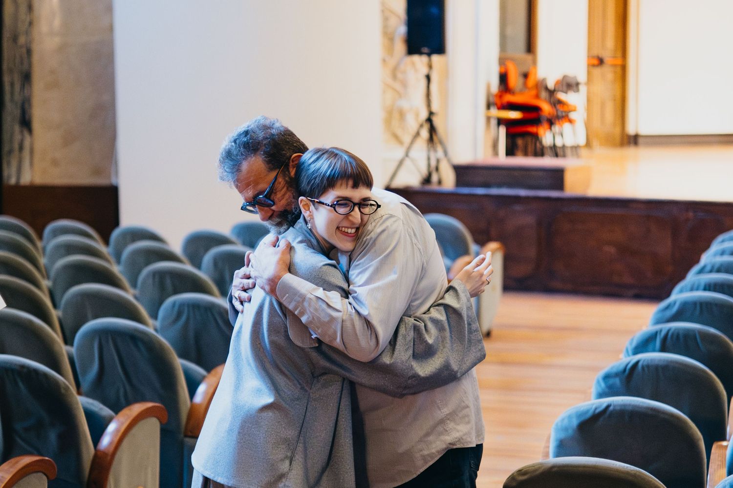 Two people share a warm hug in an elegant concert hall with rows of blue chairs.