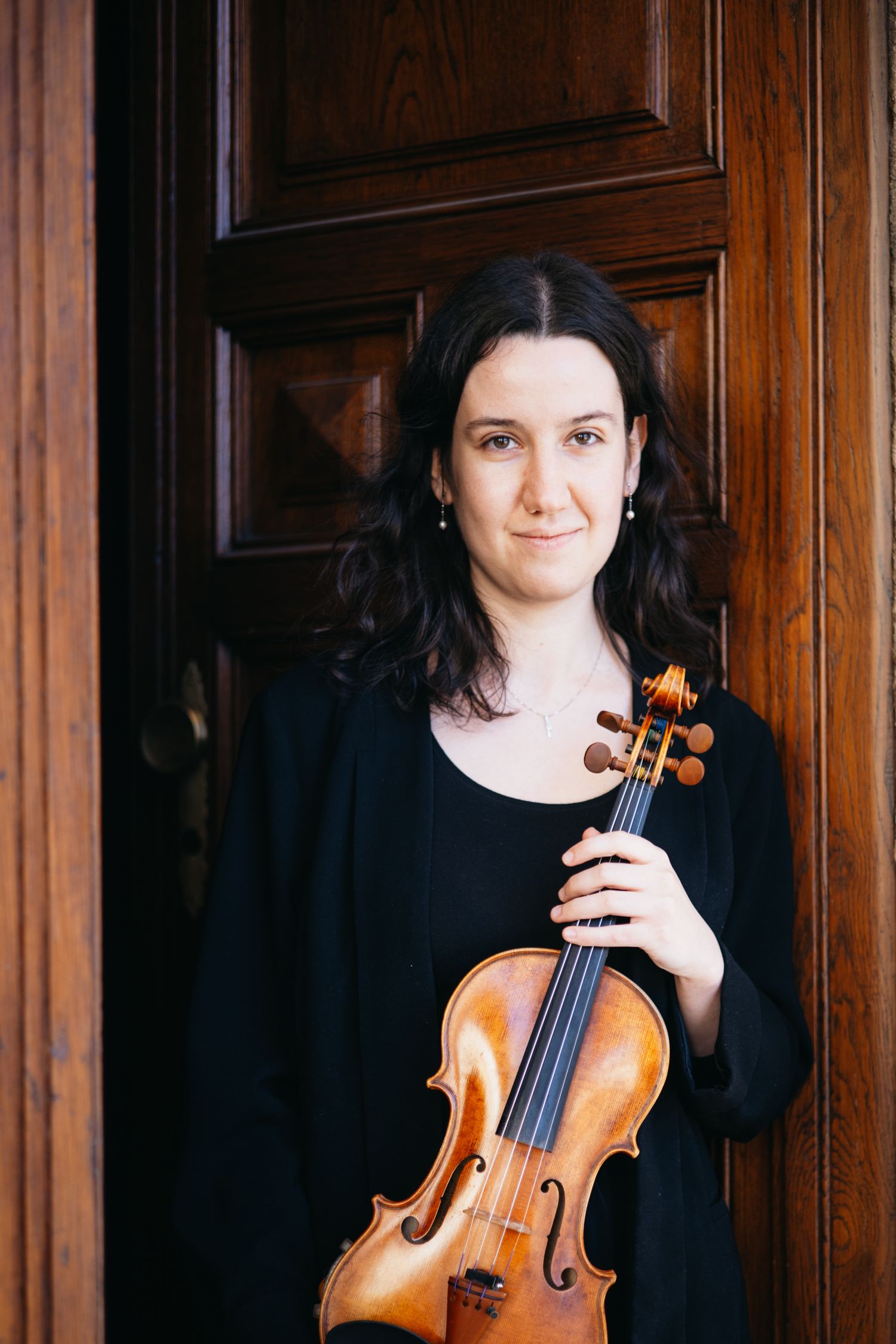 Woman in black outfit holding a violin, smiling in front of a wooden door.