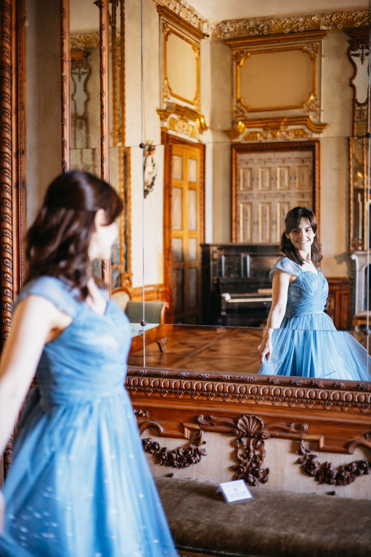 Young woman in blue ball gown admiring herself in ornate mirror inside elegant baroque room.