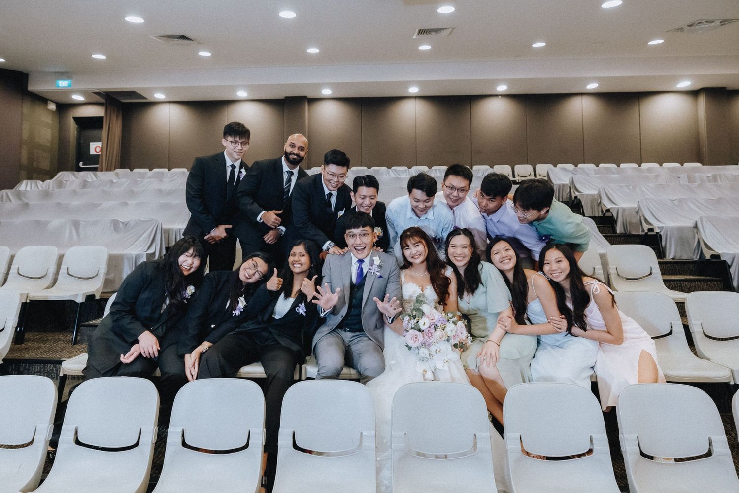 Wedding party members pose together in white auditorium chairs for a group photo.