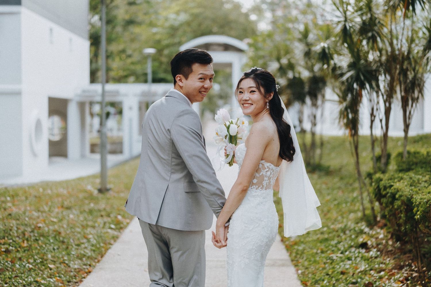 A newlywed couple walks hand in hand along a garden path in elegant wedding attire.