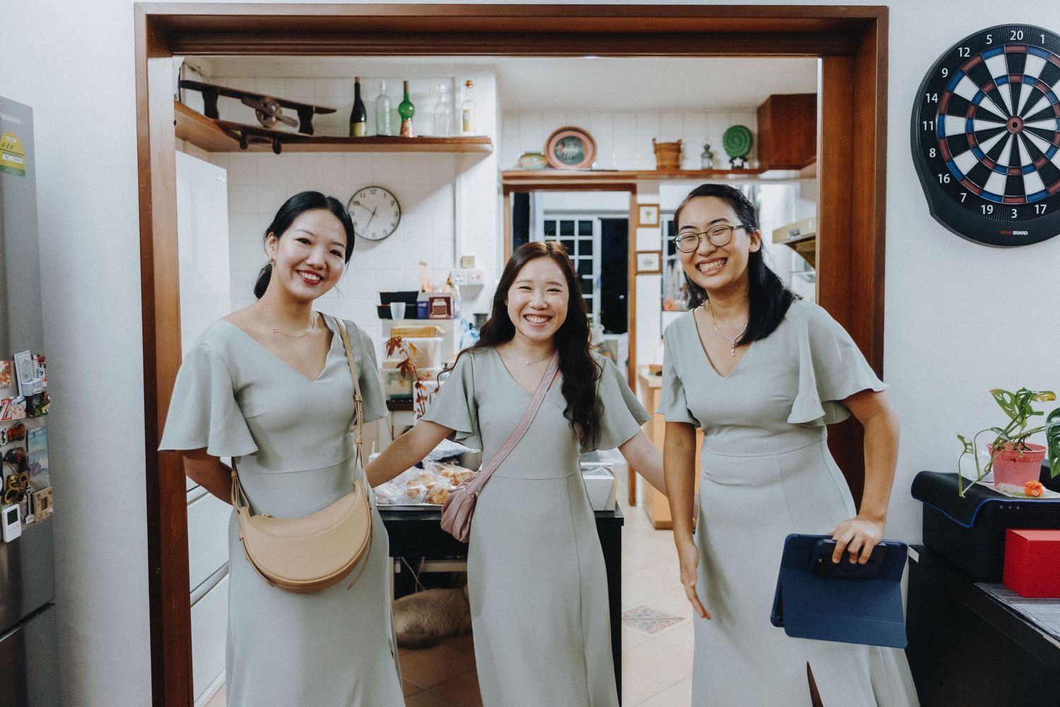 Three people in matching gray dresses pose together in a kitchen space with a dartboard on the wall.