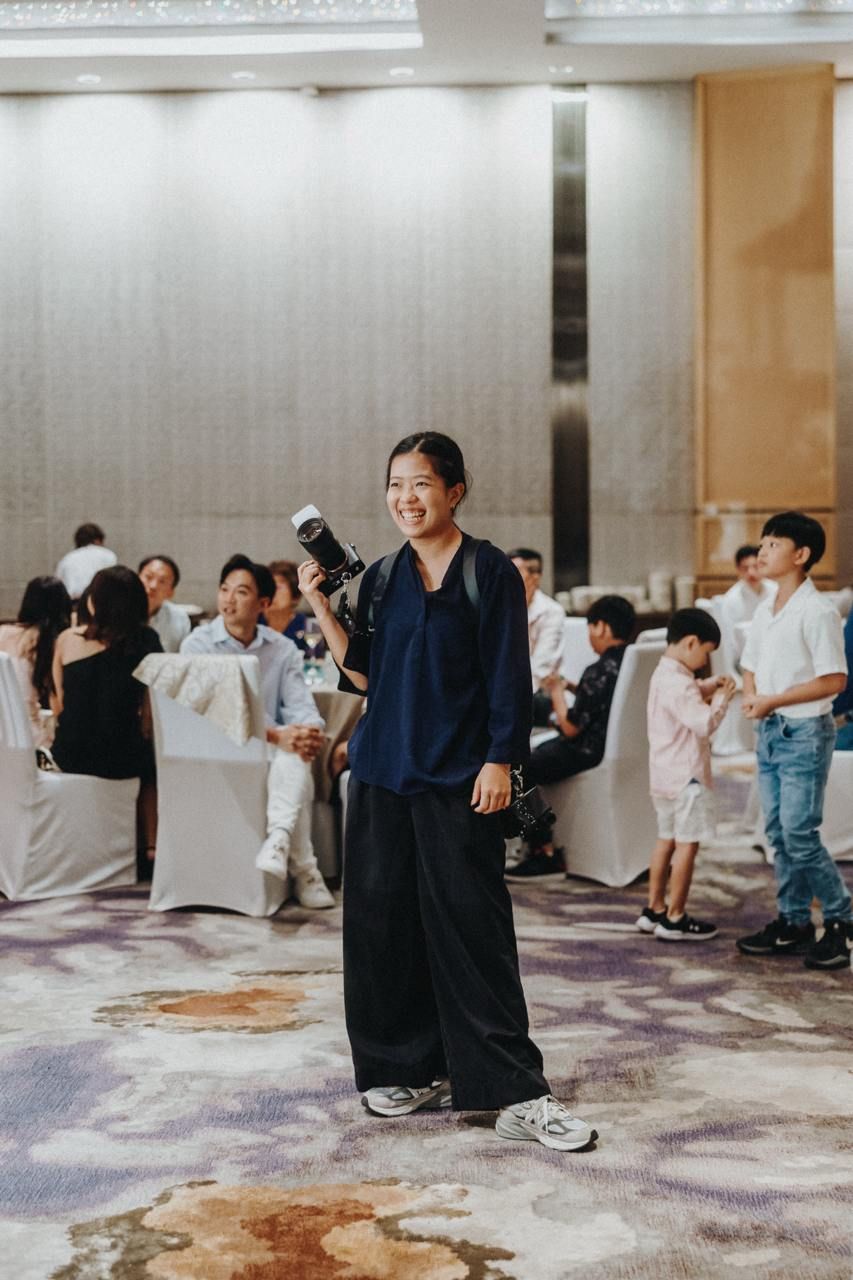 Professional photographer in navy clothing stands on patterned carpet at indoor event venue with guests in background.