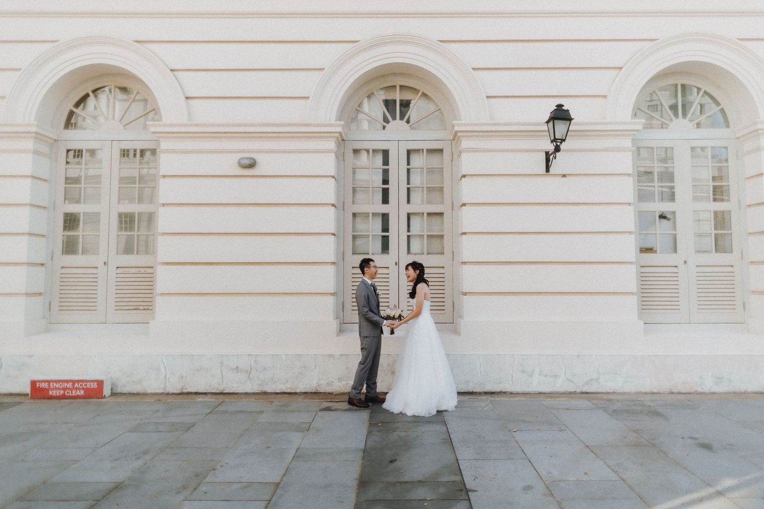 Bride and groom pose along a classic white building facade with arched windows for wedding photos.