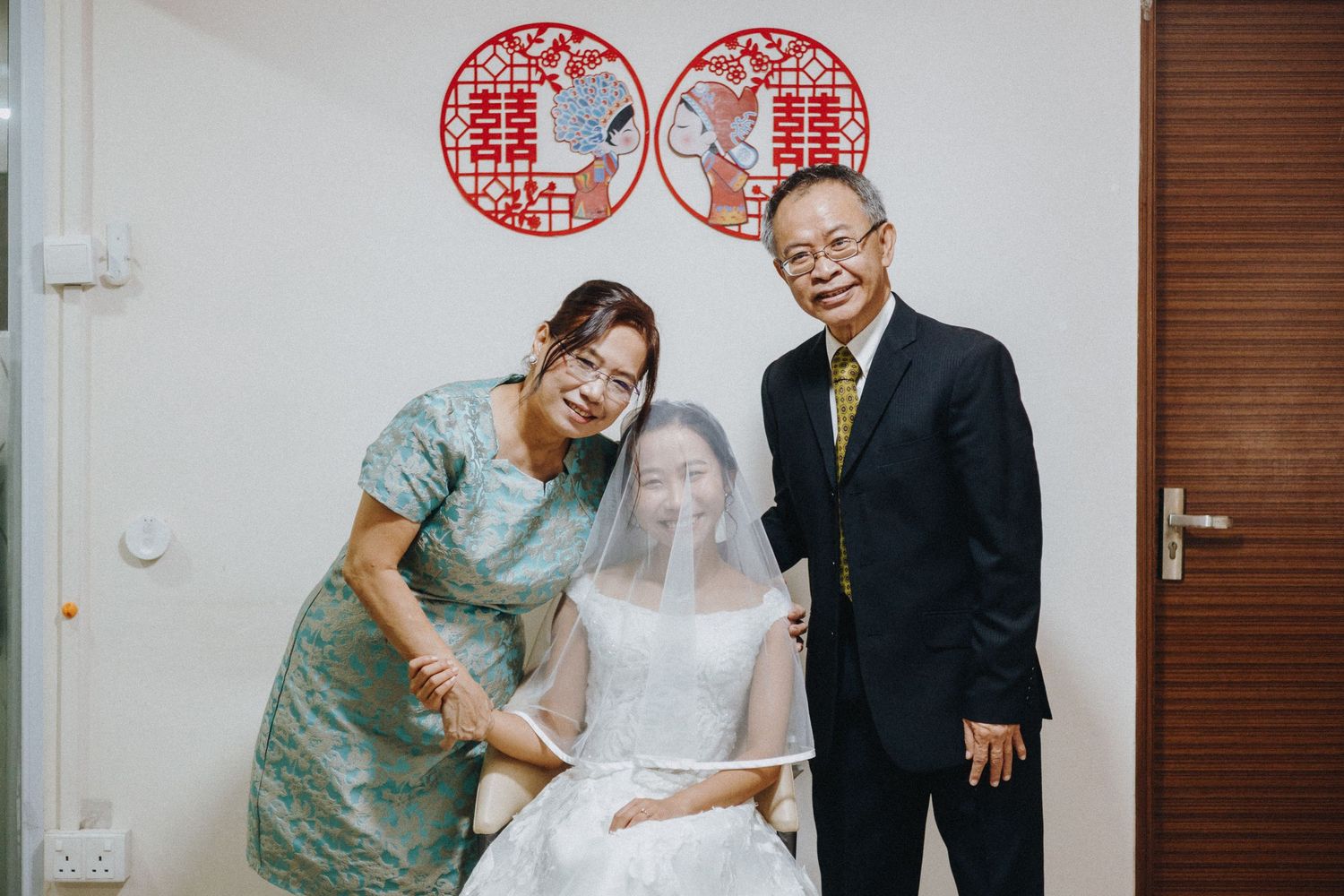 Three people pose in front of a wall with traditional red Chinese double happiness symbols for a wedding celebration.