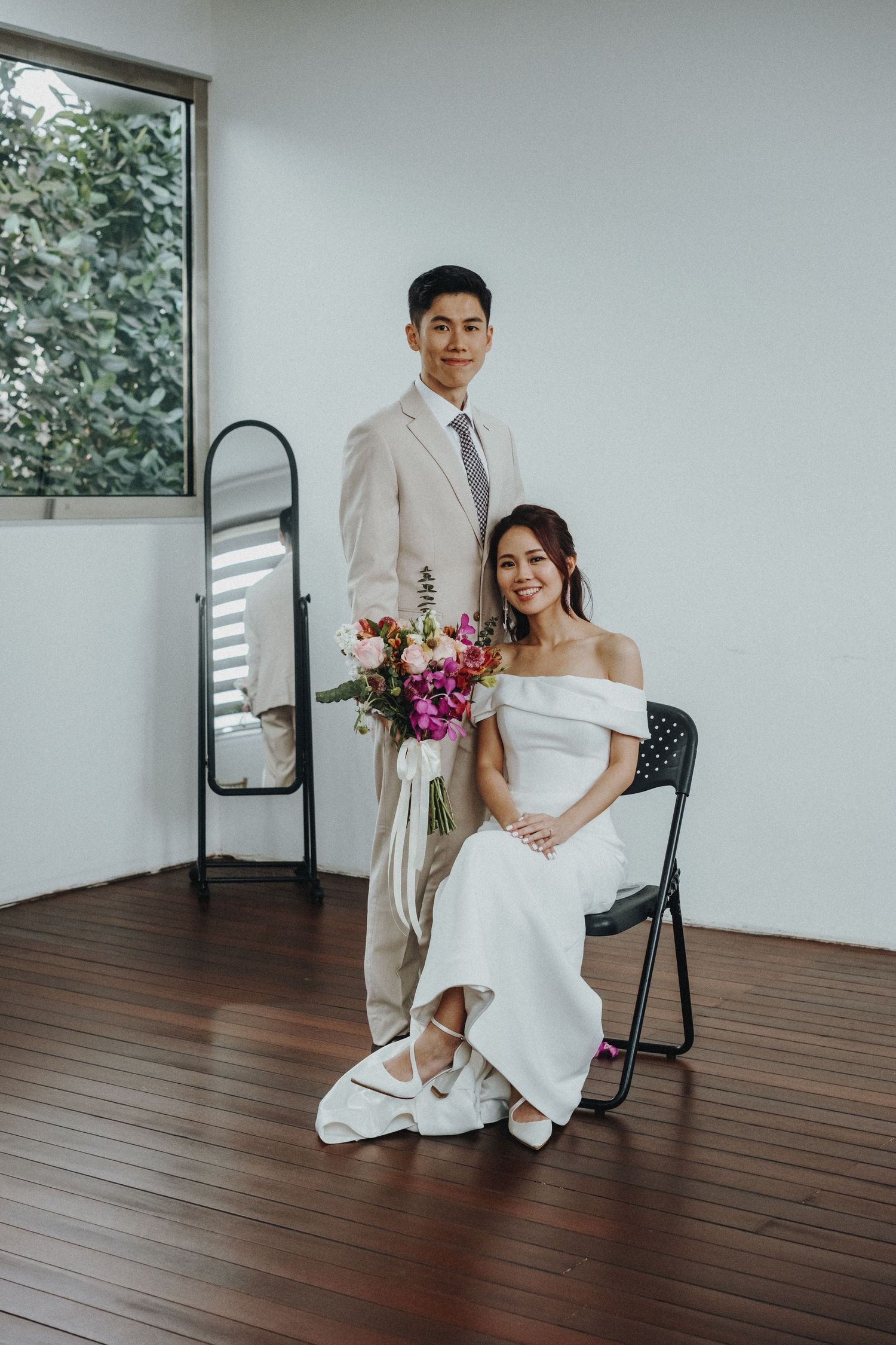 A wedding couple poses together on wooden floor against white wall in minimalist indoor setting.