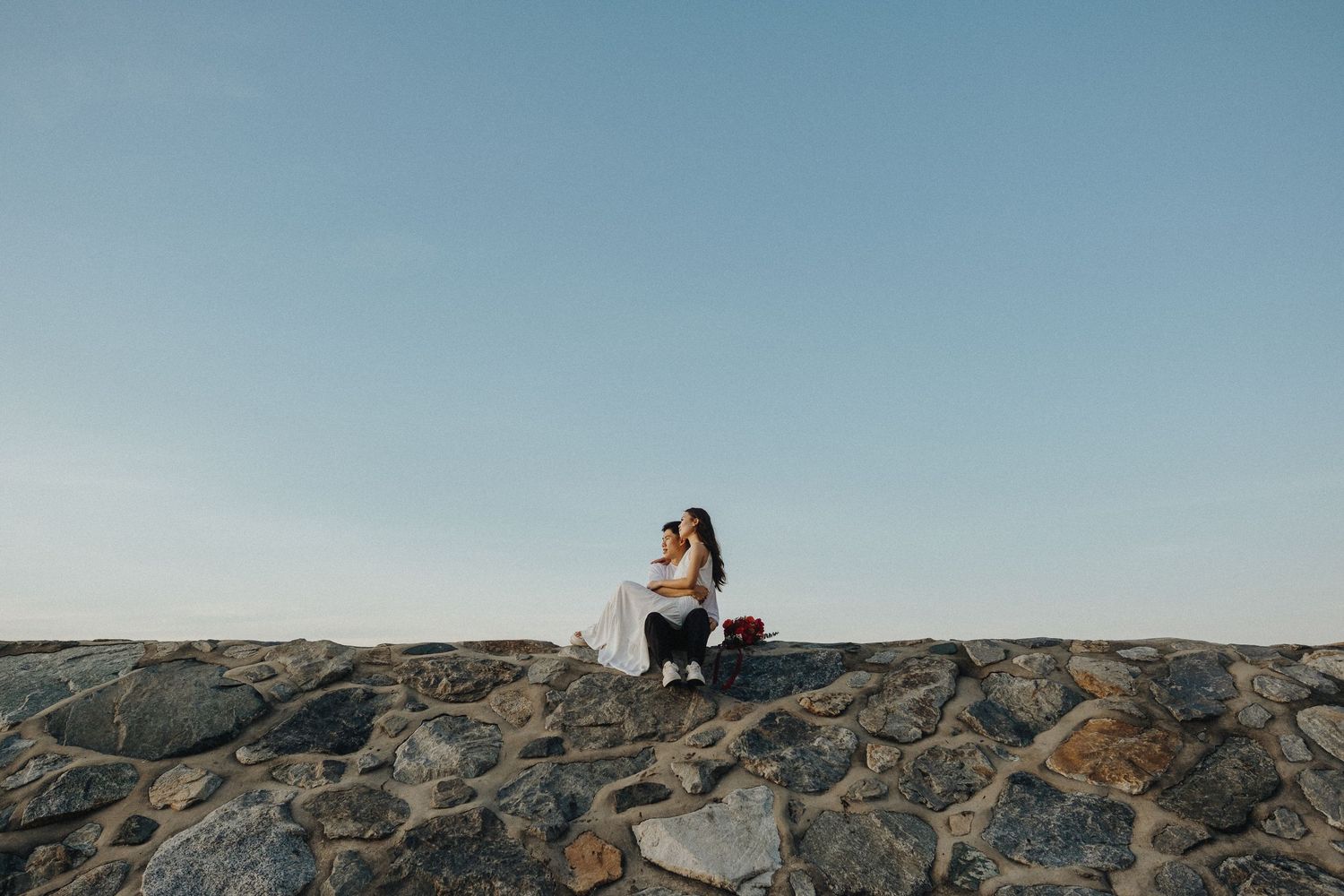 A person sits on a rocky cliff edge overlooking a scenic mountainous landscape under a clear blue sky.
