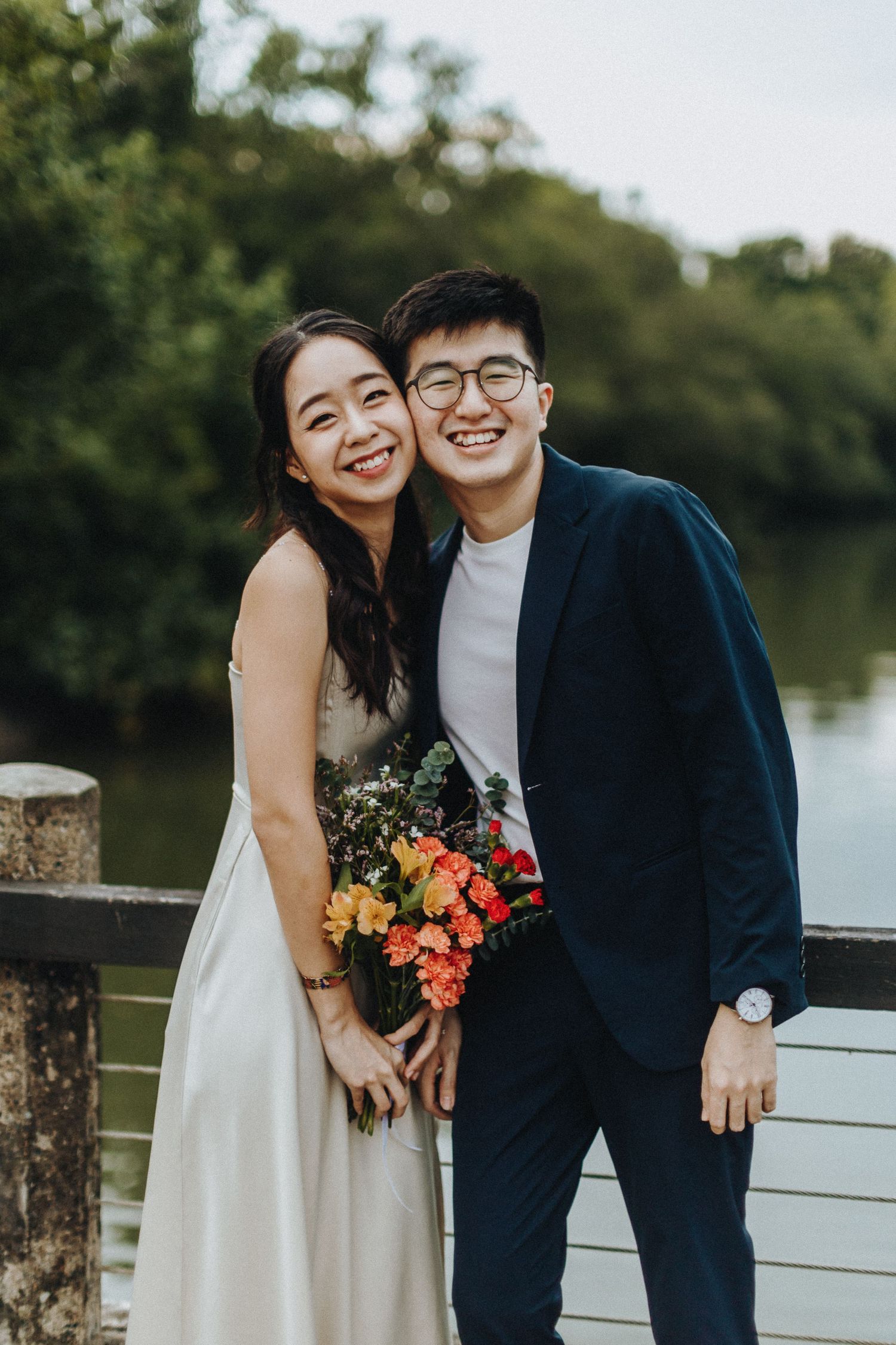 A joyful couple in wedding attire poses with a vibrant bouquet by a waterfront railing during an outdoor photoshoot.